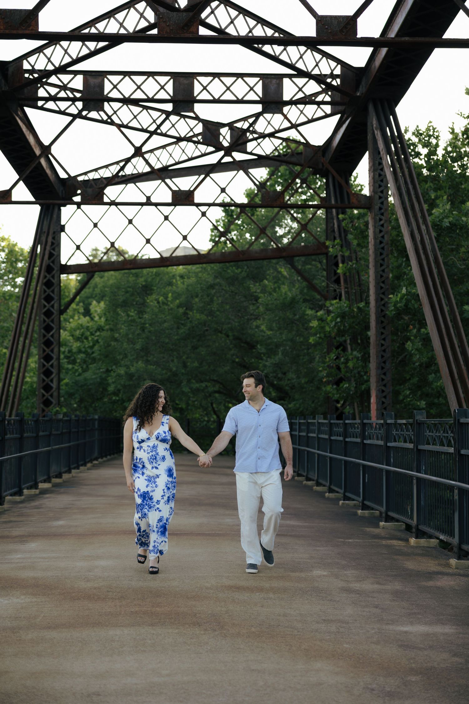 Engaged couple holding hands walking across historic iron bridge surrounded by lush green trees.