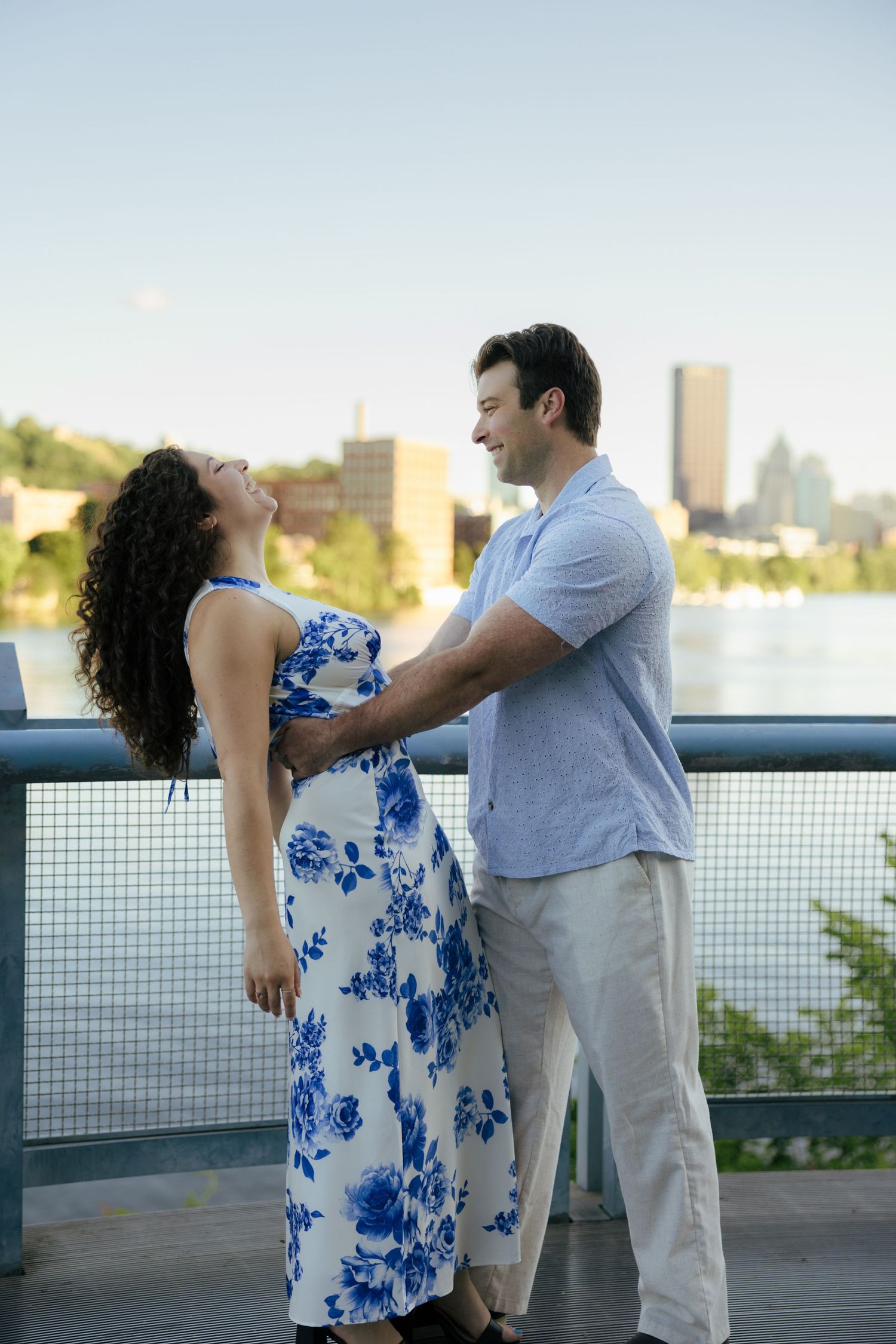 Joyful couple laughing and dancing on a bridge with a river and city skyline behind them.
