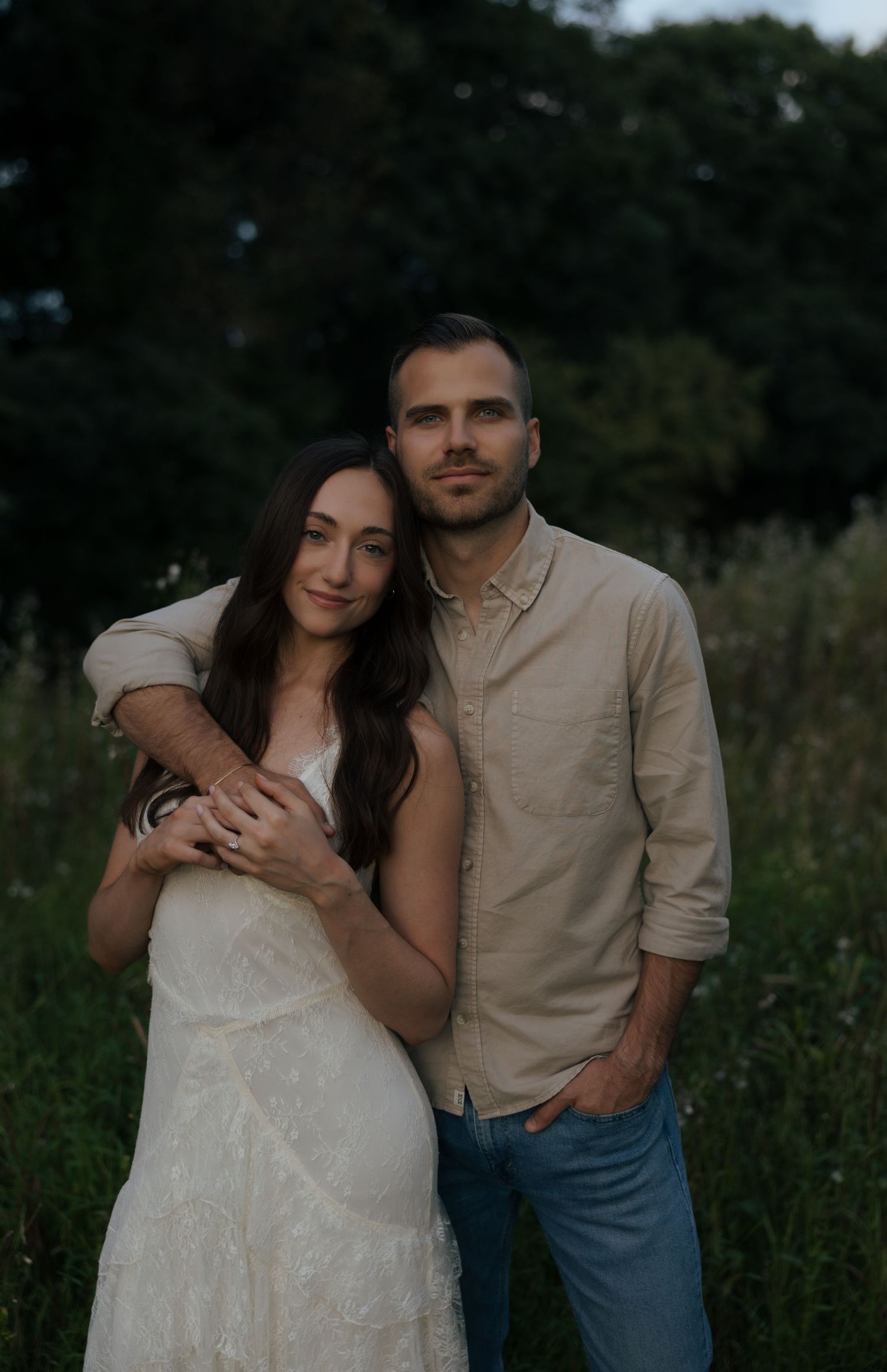 Couple embracing in a field at dusk, woman in white dress leaning against man in beige shirt and jeans.