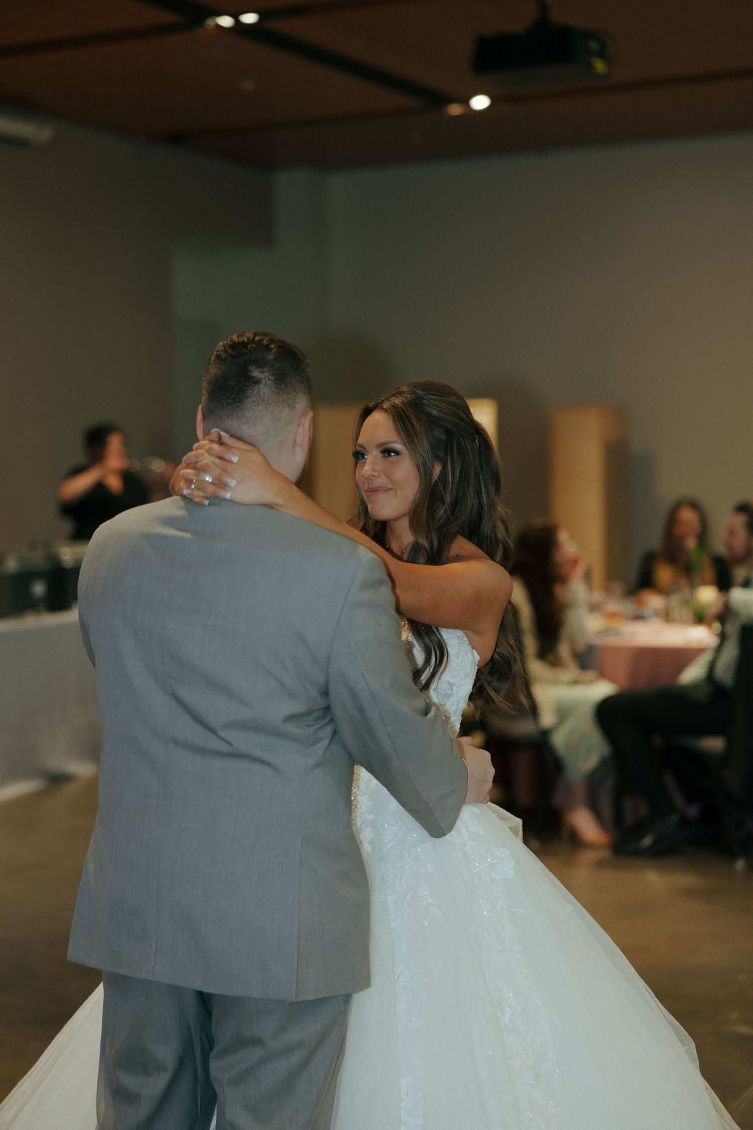 Bride in white lace gown smiles warmly at groom during first dance at elegant indoor wedding reception.