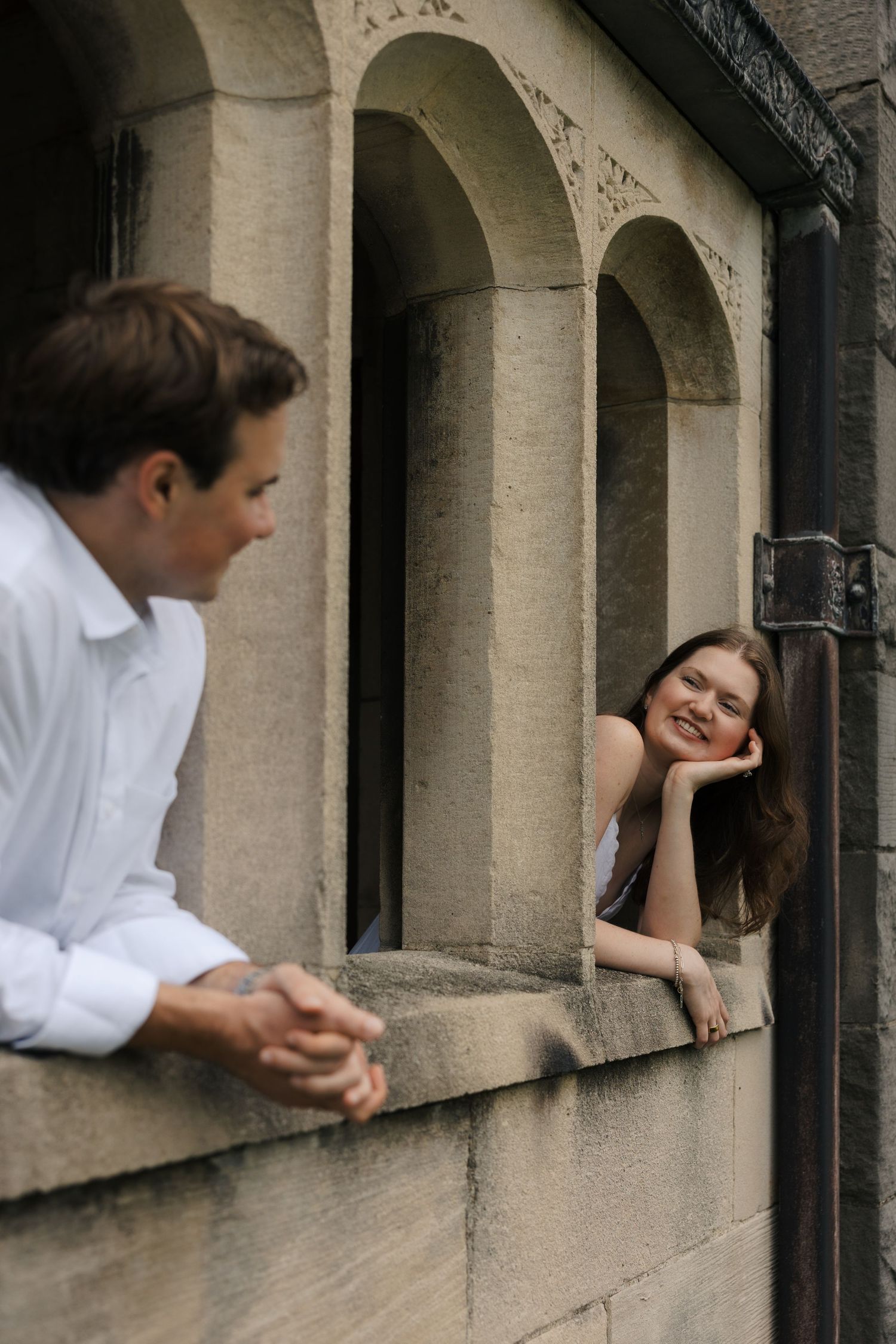 Woman leaning on stone window ledge smiling at man standing outside a historic stone building.