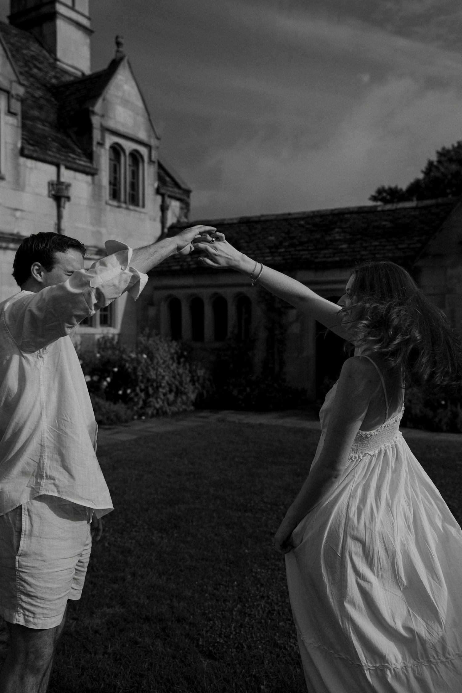 Black and white photo of a couple dancing outdoors, holding hands raised, near a historic stone building.
