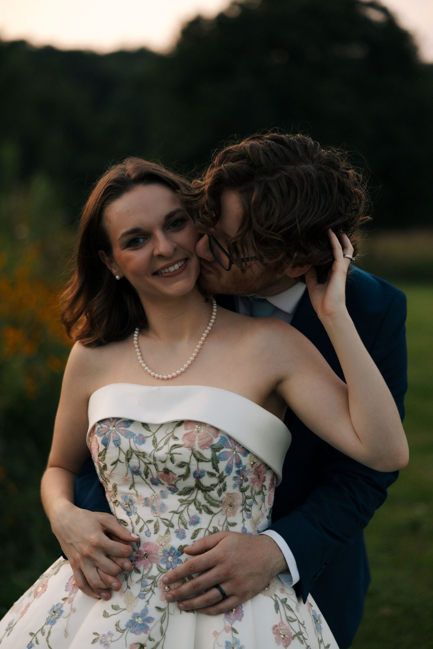 Groom in navy suit kisses bride on cheek as she smiles wearing a strapless floral gown and pearl necklace.