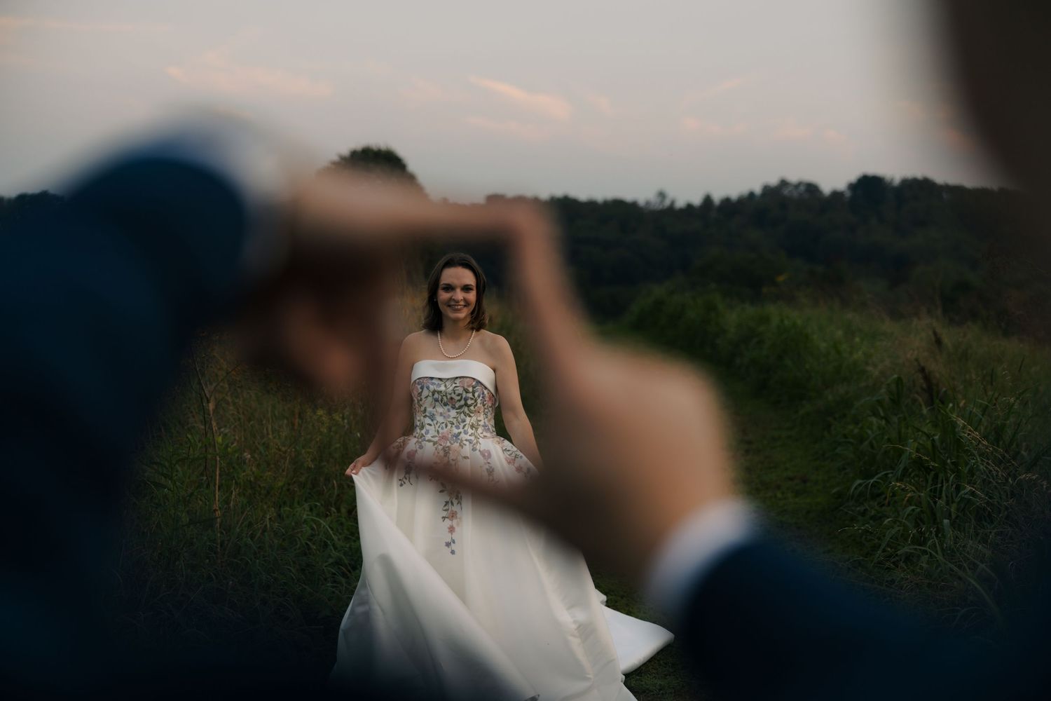 Groom frames bride in white floral wedding gown through his fingers in a lush green field.