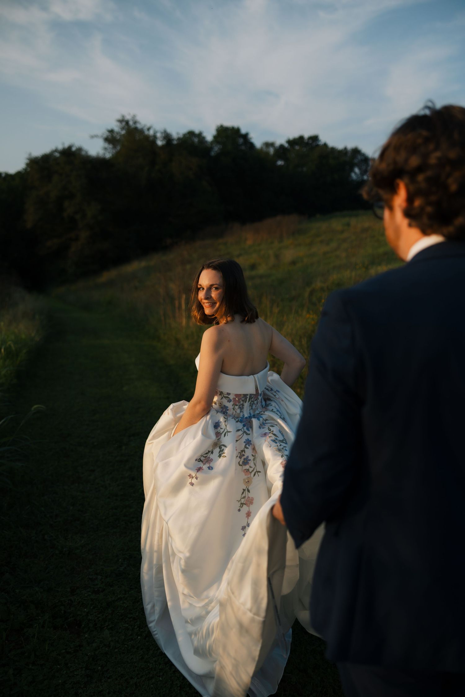 Bride in floral embroidered white gown looks back smiling at groom in navy suit during golden hour outdoor wedding.