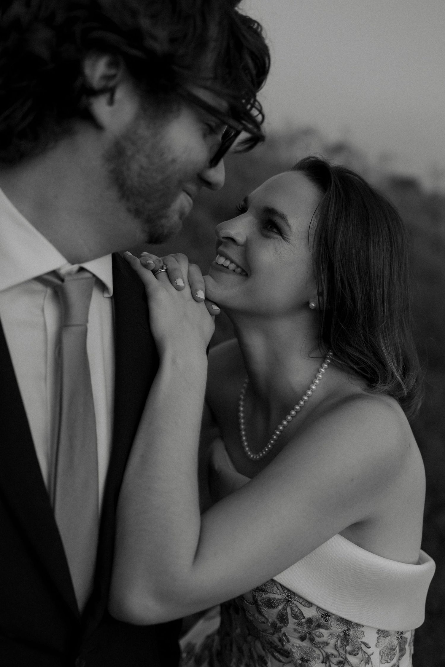 Black and white close-up of smiling bride in pearls gazing up at groom holding his lapel.