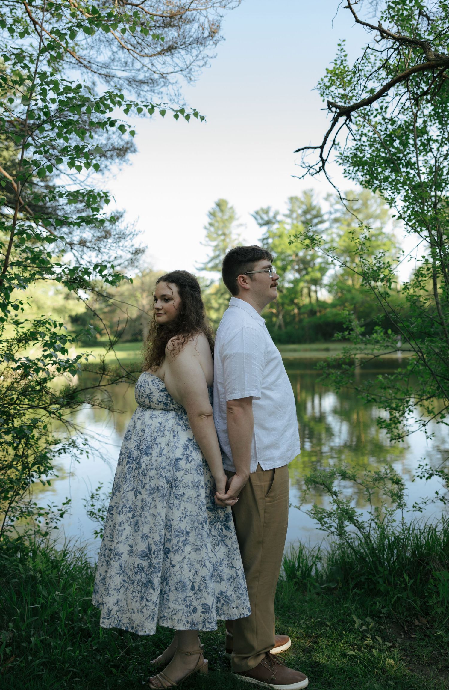 Engaged couple stands back-to-back holding hands by a reflective pond, woman in blue floral dress smiles softly.