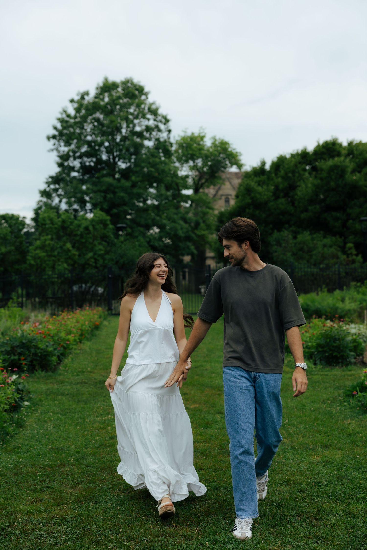 Happy couple walking hand in hand through a green garden, woman in flowing white dress laughing with her partner.