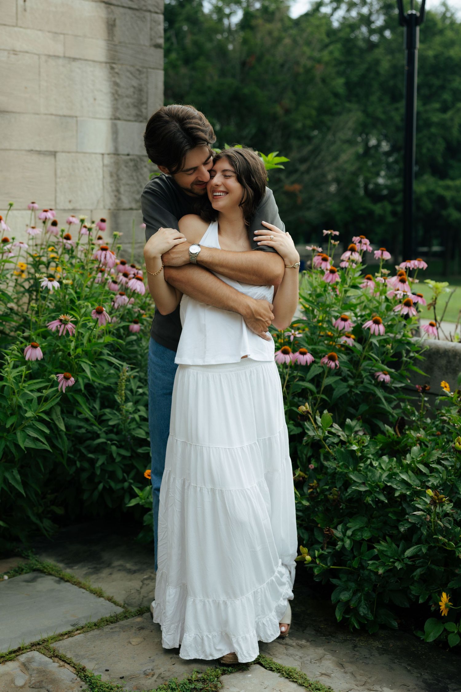 A couple embraces near a stone wall surrounded by blooming pink echinacea flowers in a garden.