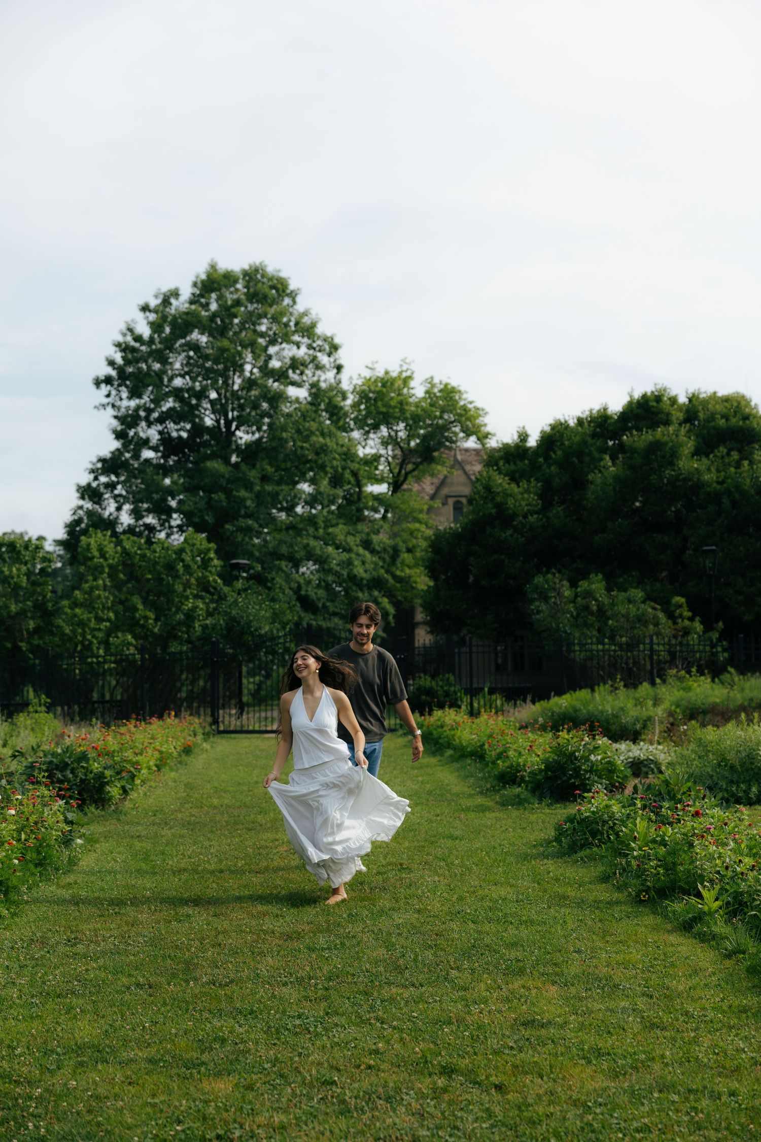 Couple running together through a lush garden path, woman in white dress, man in dark shirt, surrounded by greenery.