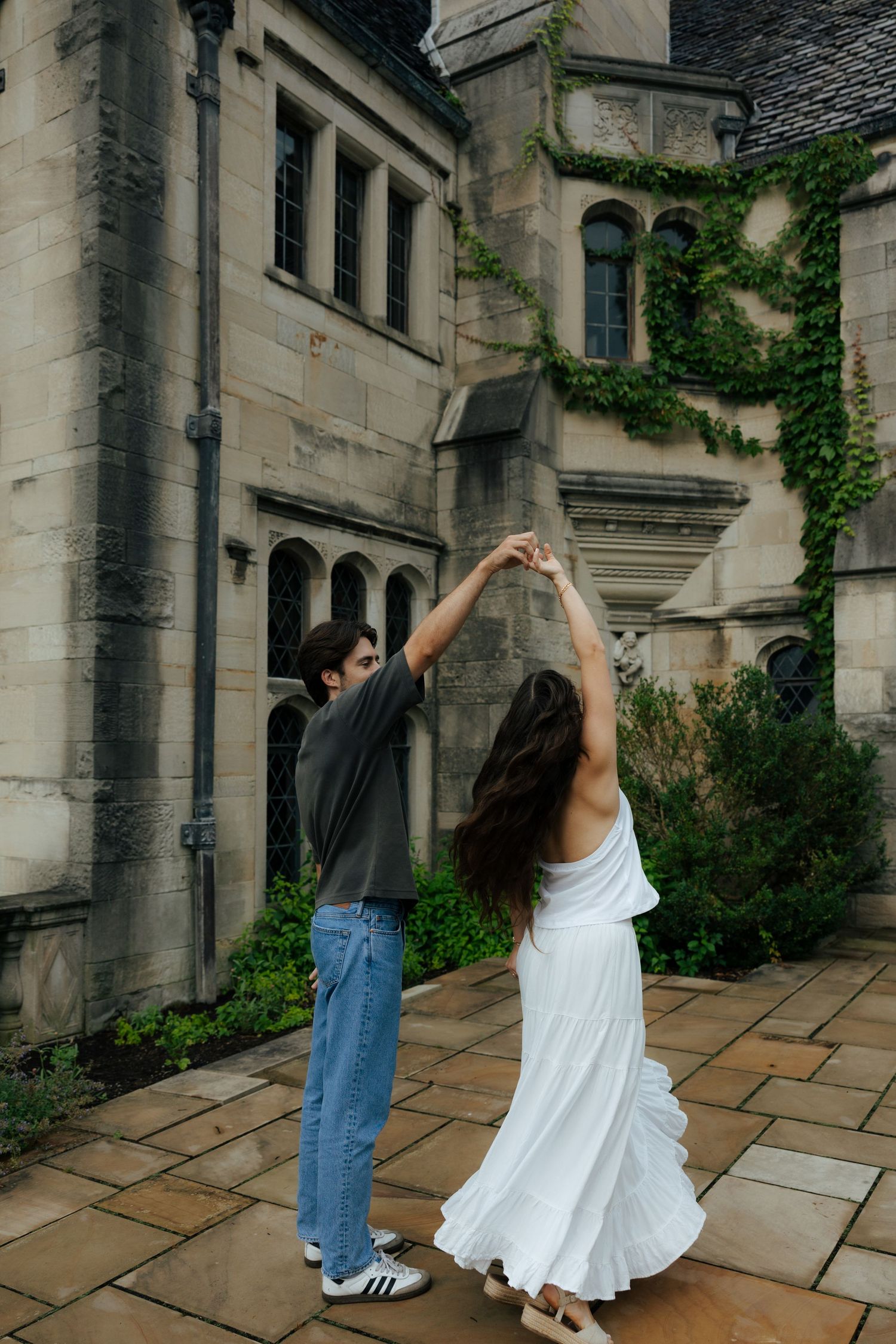Couple dancing outdoors near a stone mansion covered in ivy, woman in flowing white dress.
