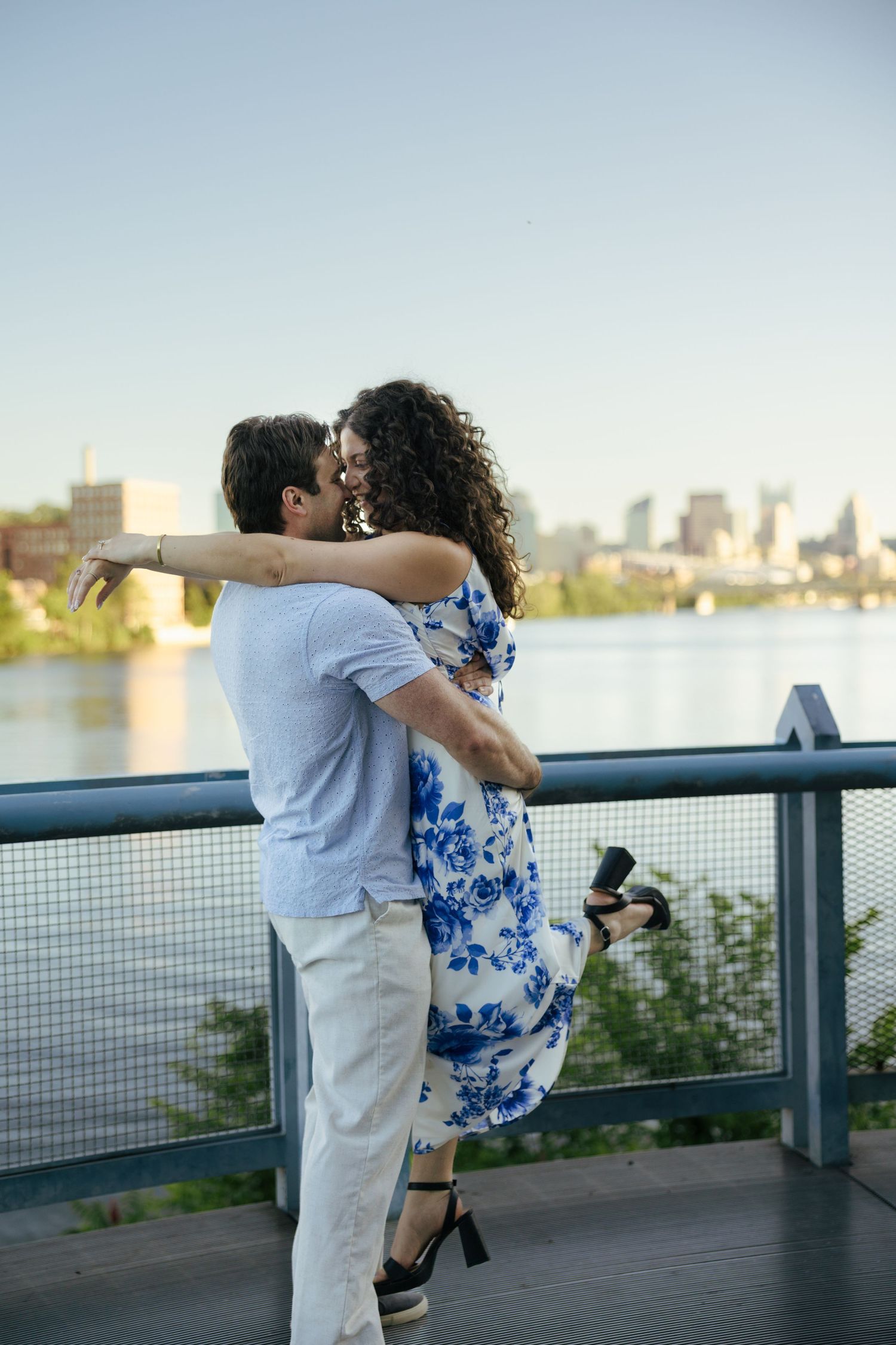 Couple embracing and kissing on a waterfront bridge with a city skyline in the background.