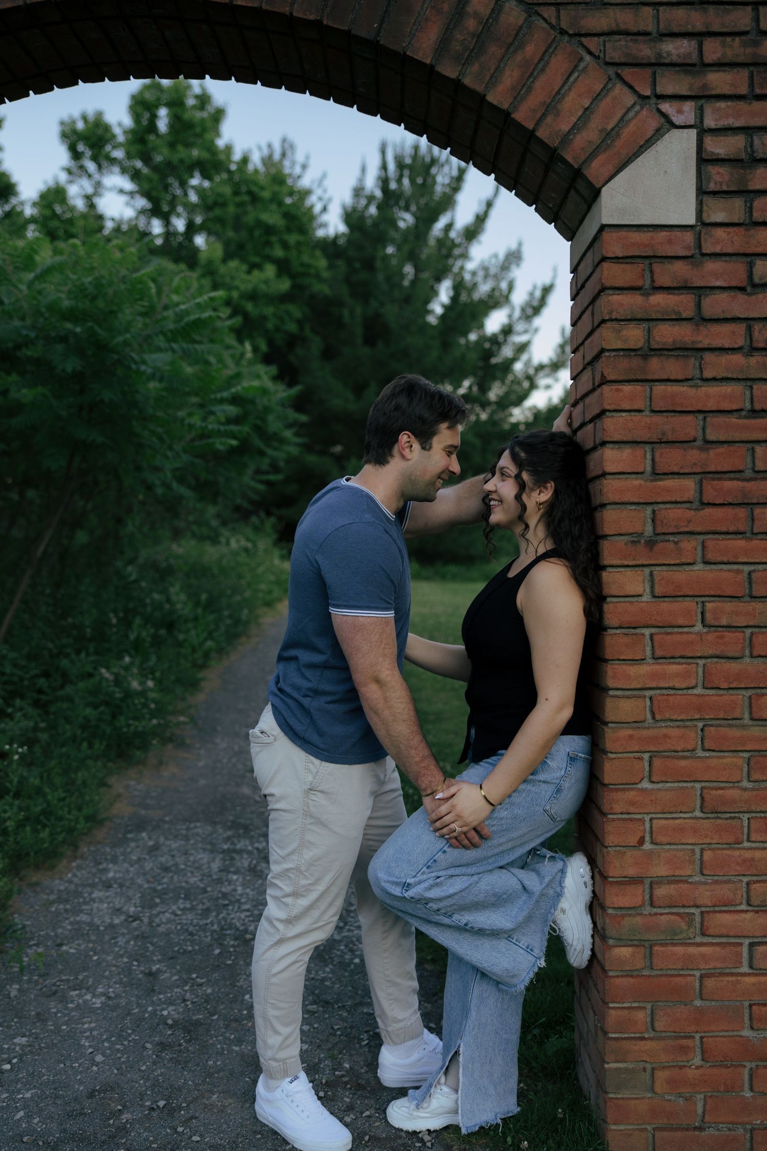 Romantic couple holding hands and leaning close under a brick archway surrounded by lush green trees.