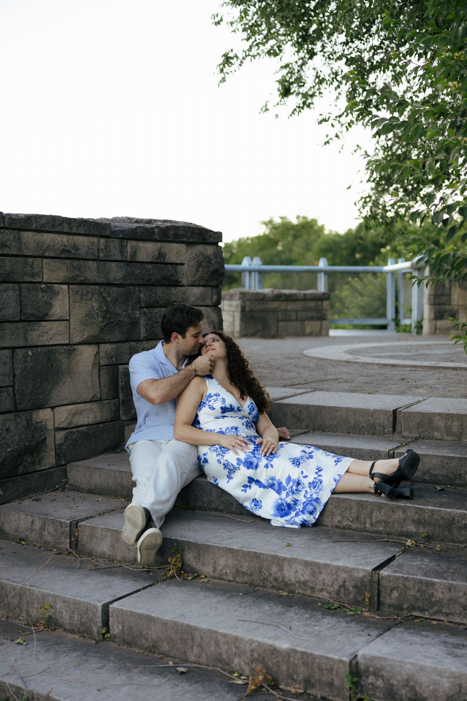 Romantic couple sitting on stone steps outdoors, man kissing womans hand in blue floral dress.