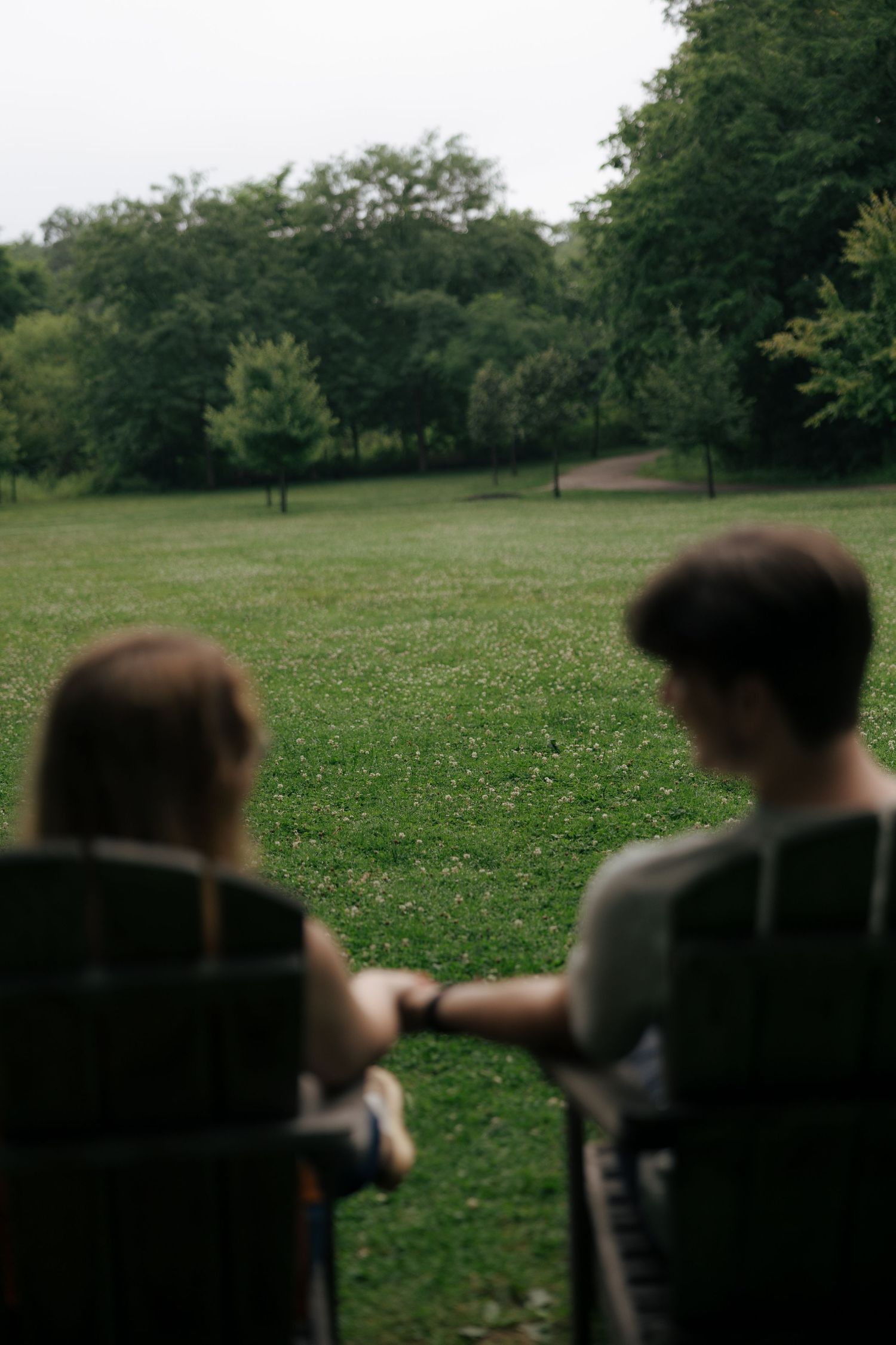 Defocused couple holding hands in wooden chairs while gazing at a lush green park landscape.