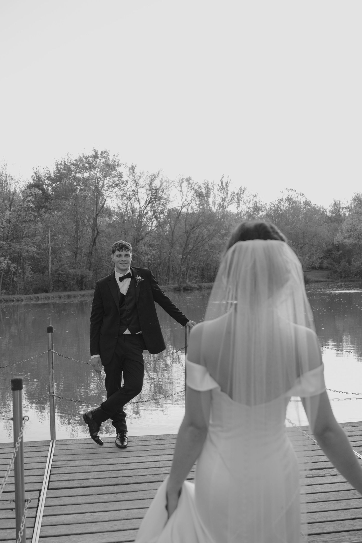 Black and white photo of bride walking toward groom on wooden dock beside calm reflective lake surrounded by trees.