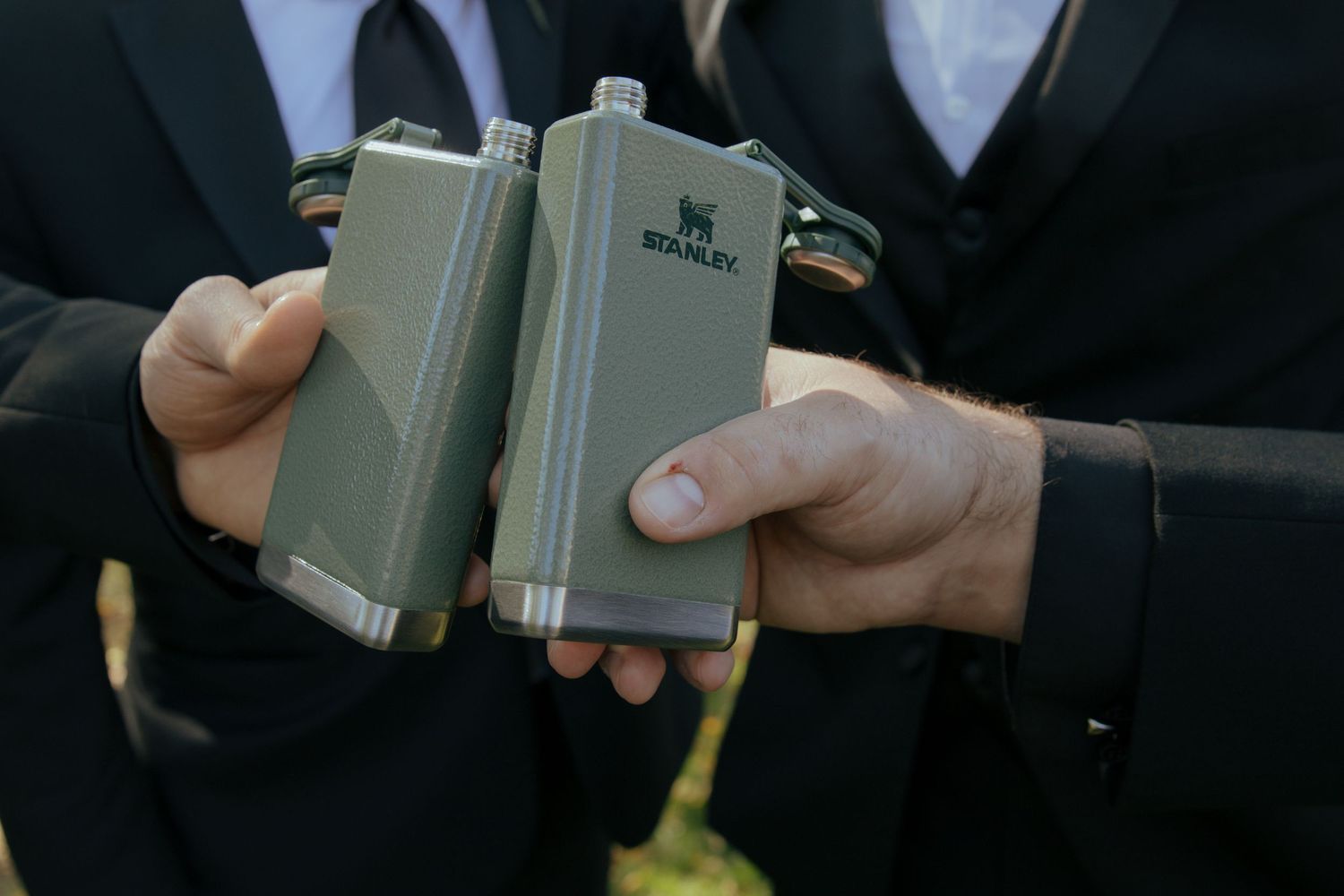 Two groomsmen in black suits toasting with Stanley brand olive green hip flasks outdoors.