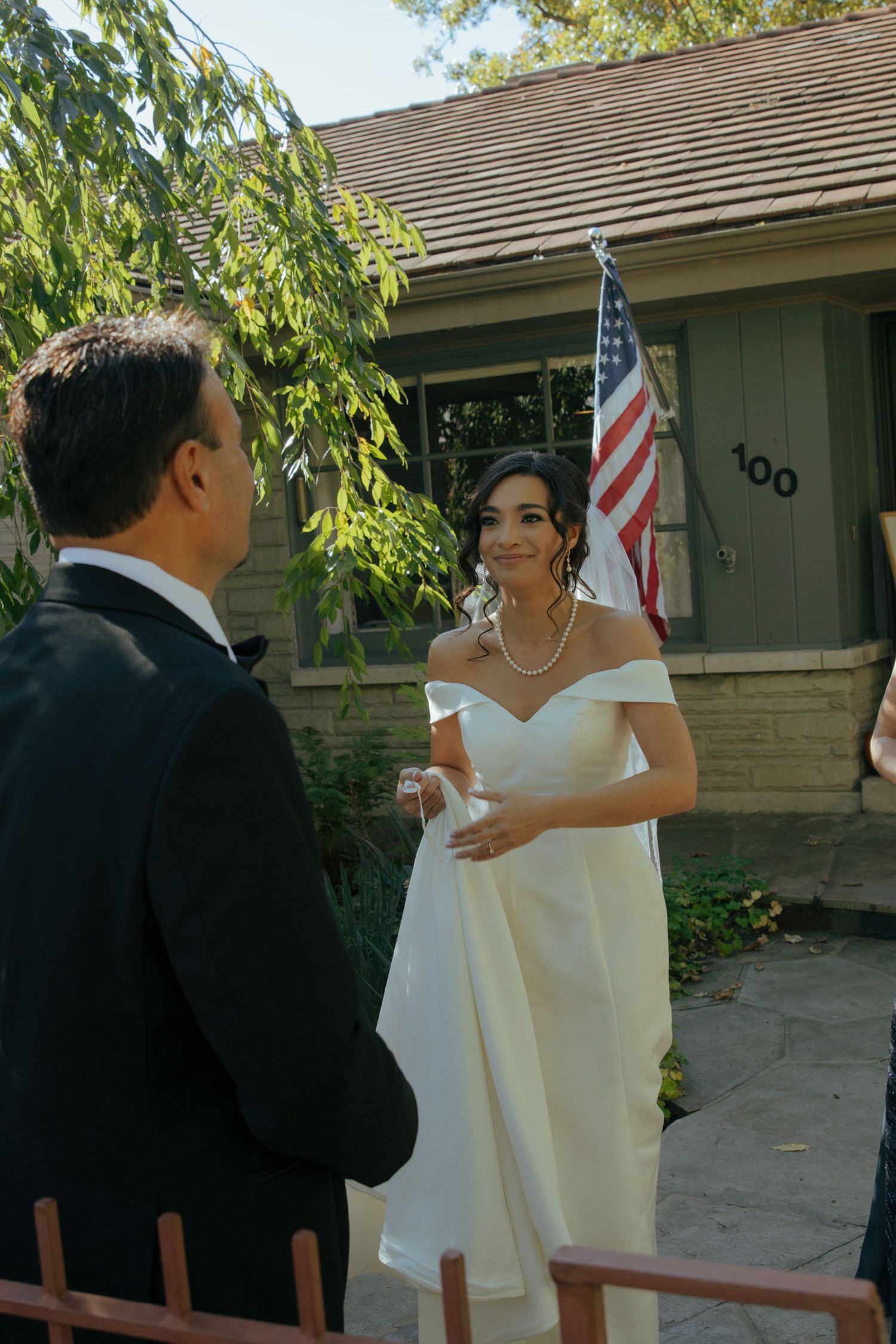 Bride in white off-shoulder gown exchanges vows with groom outdoors near American flag at intimate wedding ceremony.