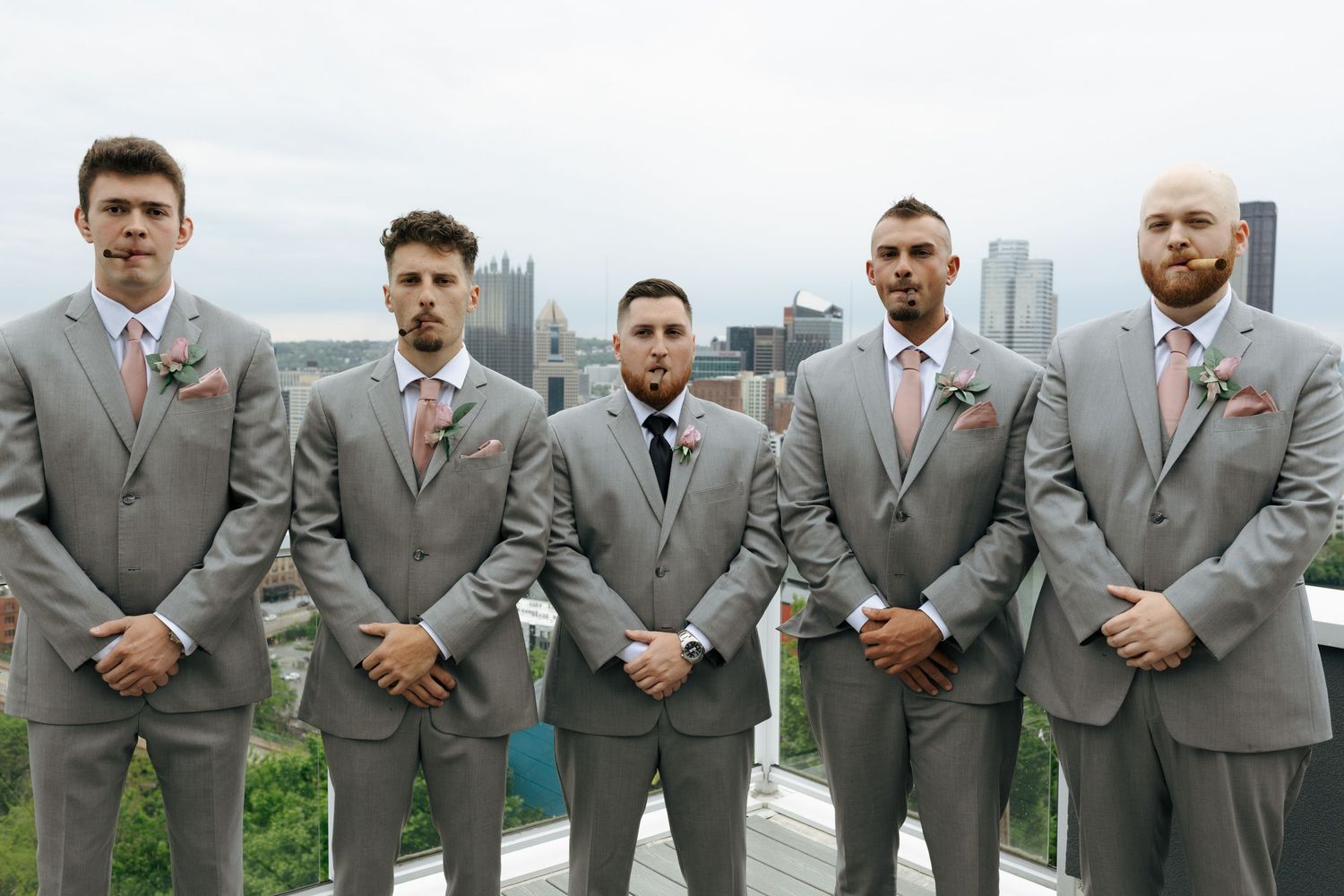 Five groomsmen in gray suits with pink boutonnieres pose outdoors with a city skyline backdrop.