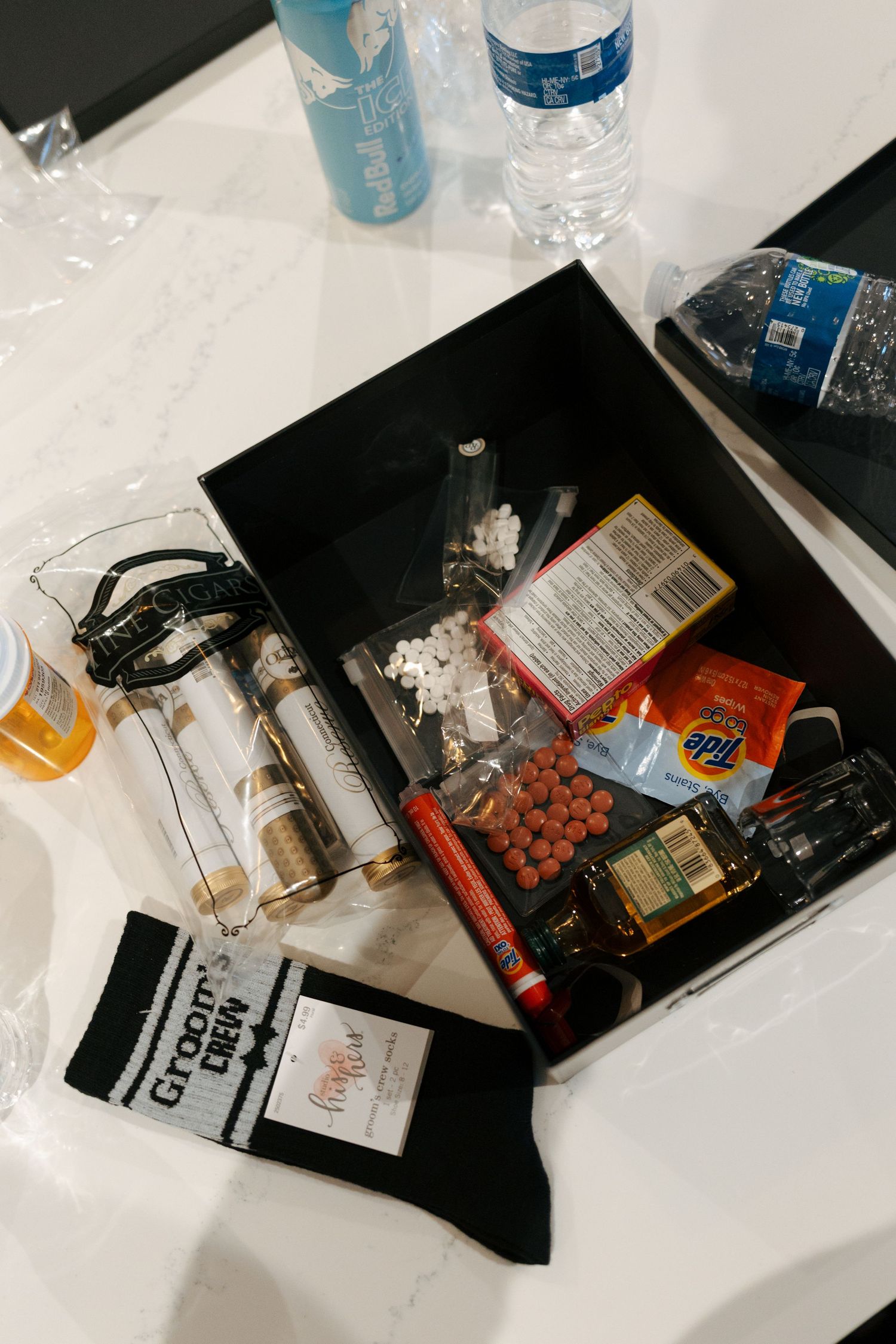 Black box filled with assorted items including socks, snacks, Tide pods, and bottles on a marble surface.
