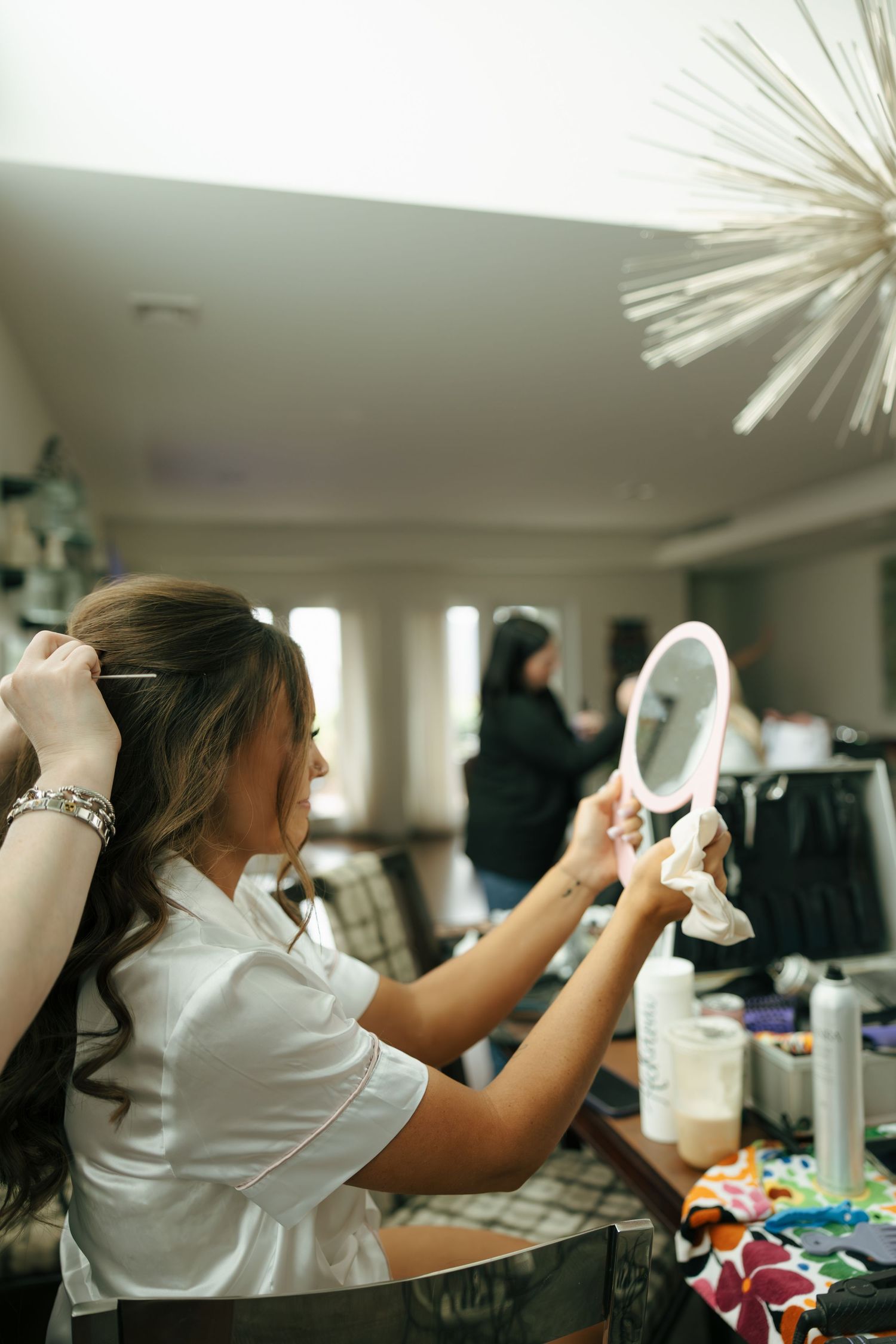 A woman styles her hair while holding a lighted mirror at a vanity table during wedding preparations.