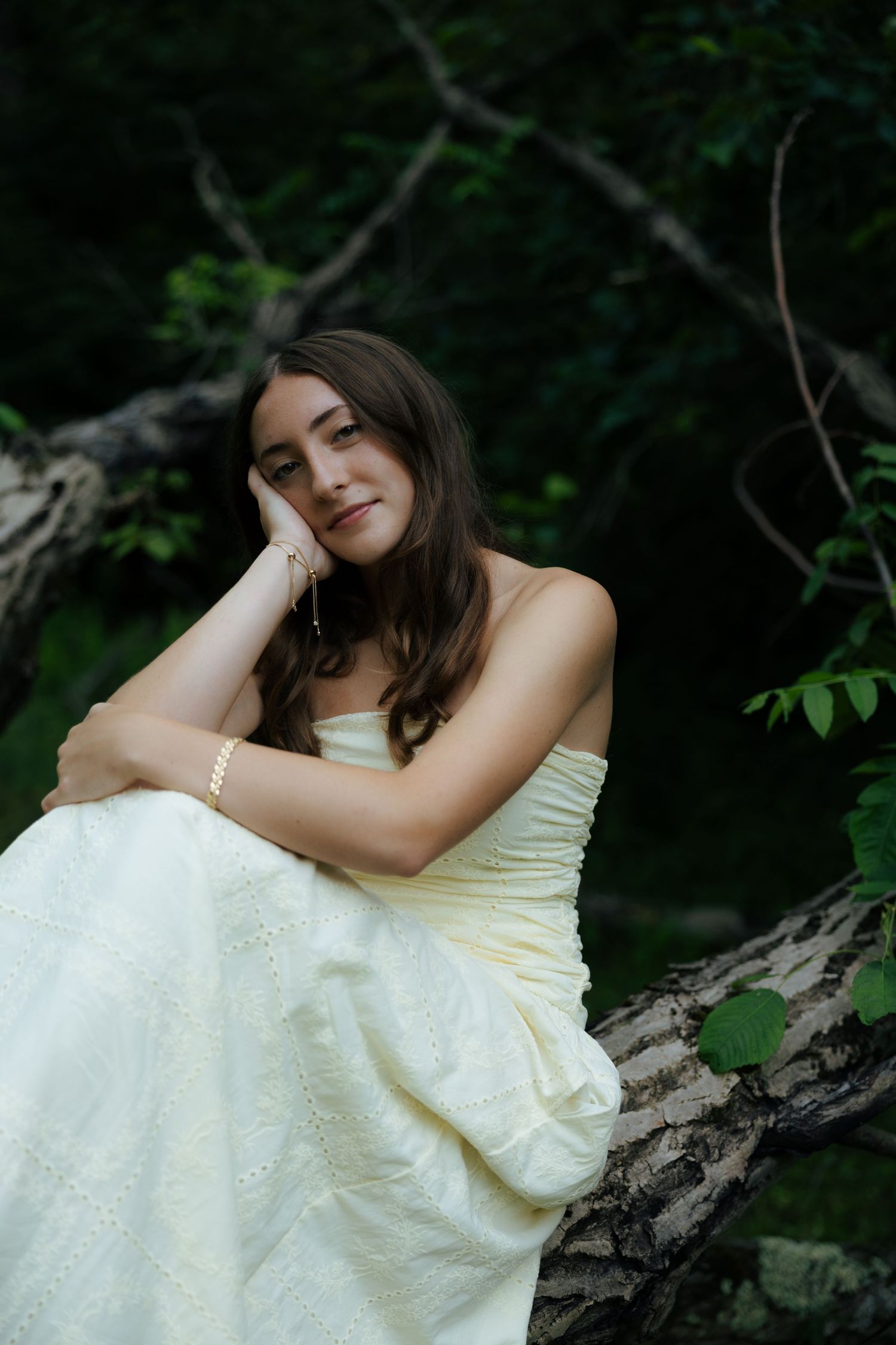 Young woman in white strapless gown resting on fallen tree in dark forest.