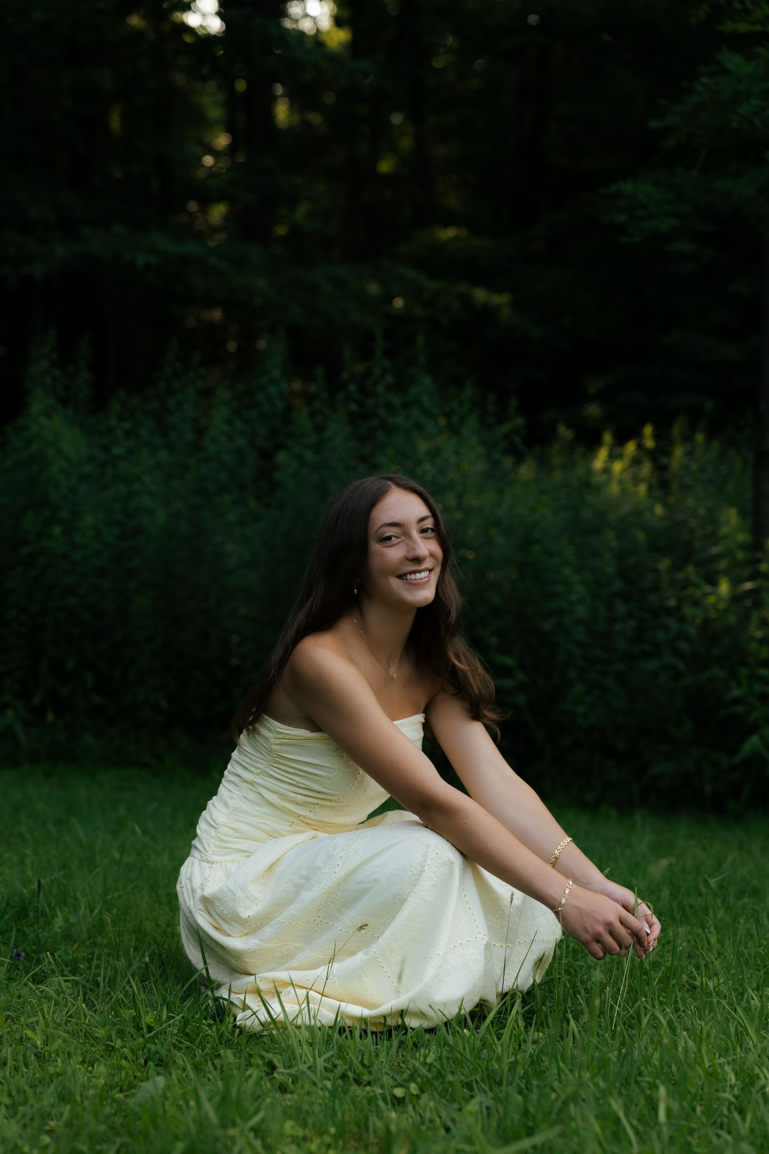A smiling young woman in a strapless white dress sits gracefully on green grass with lush trees in the background.