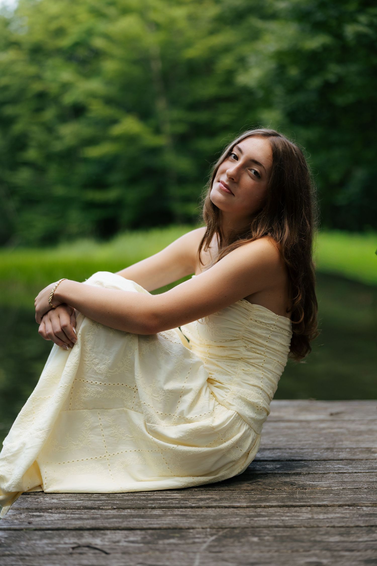 Young woman in yellow strapless dress sitting on wooden dock with green forest background.