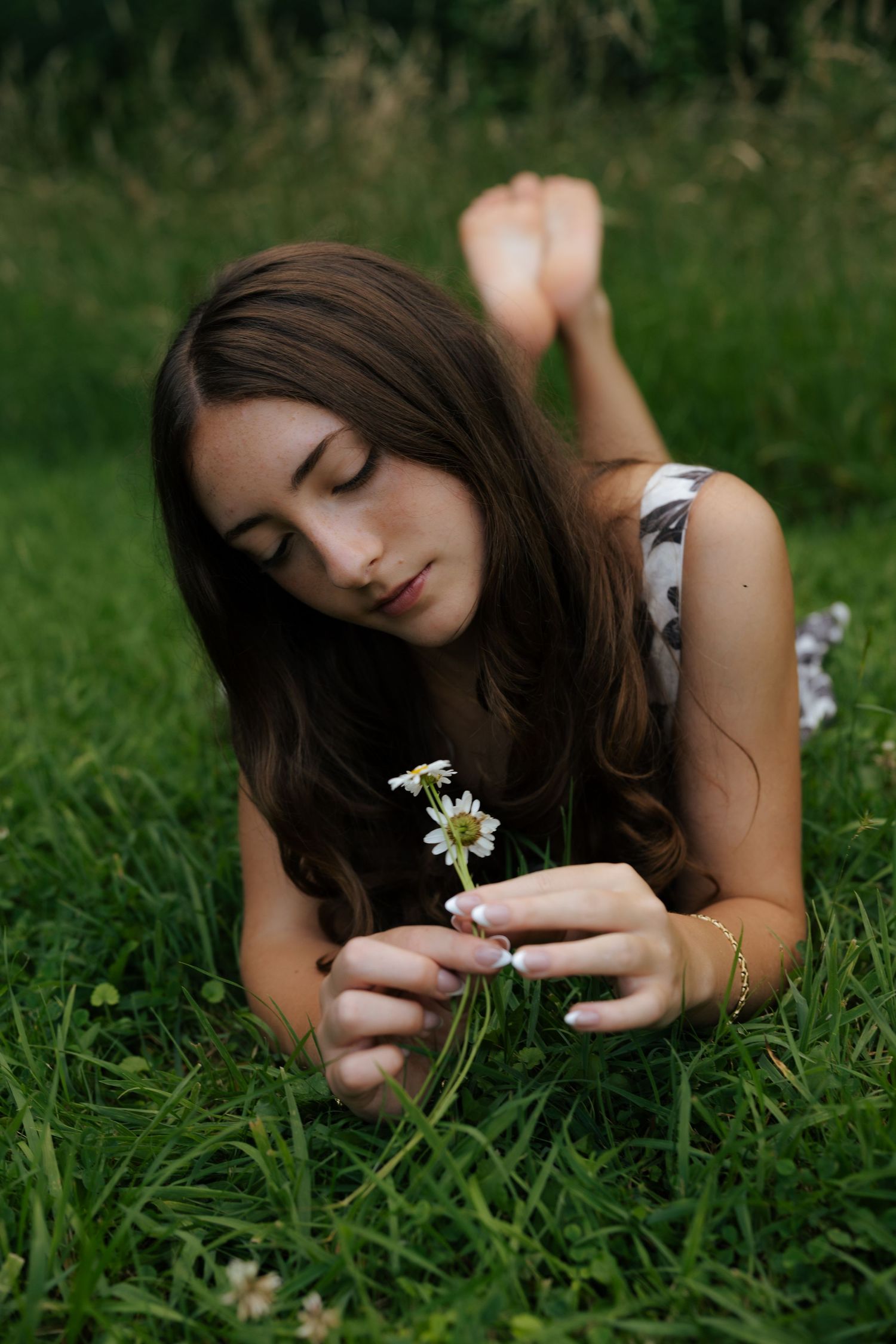 Young woman with long brown hair lying in green grass holding small white daisy flowers outdoors.
