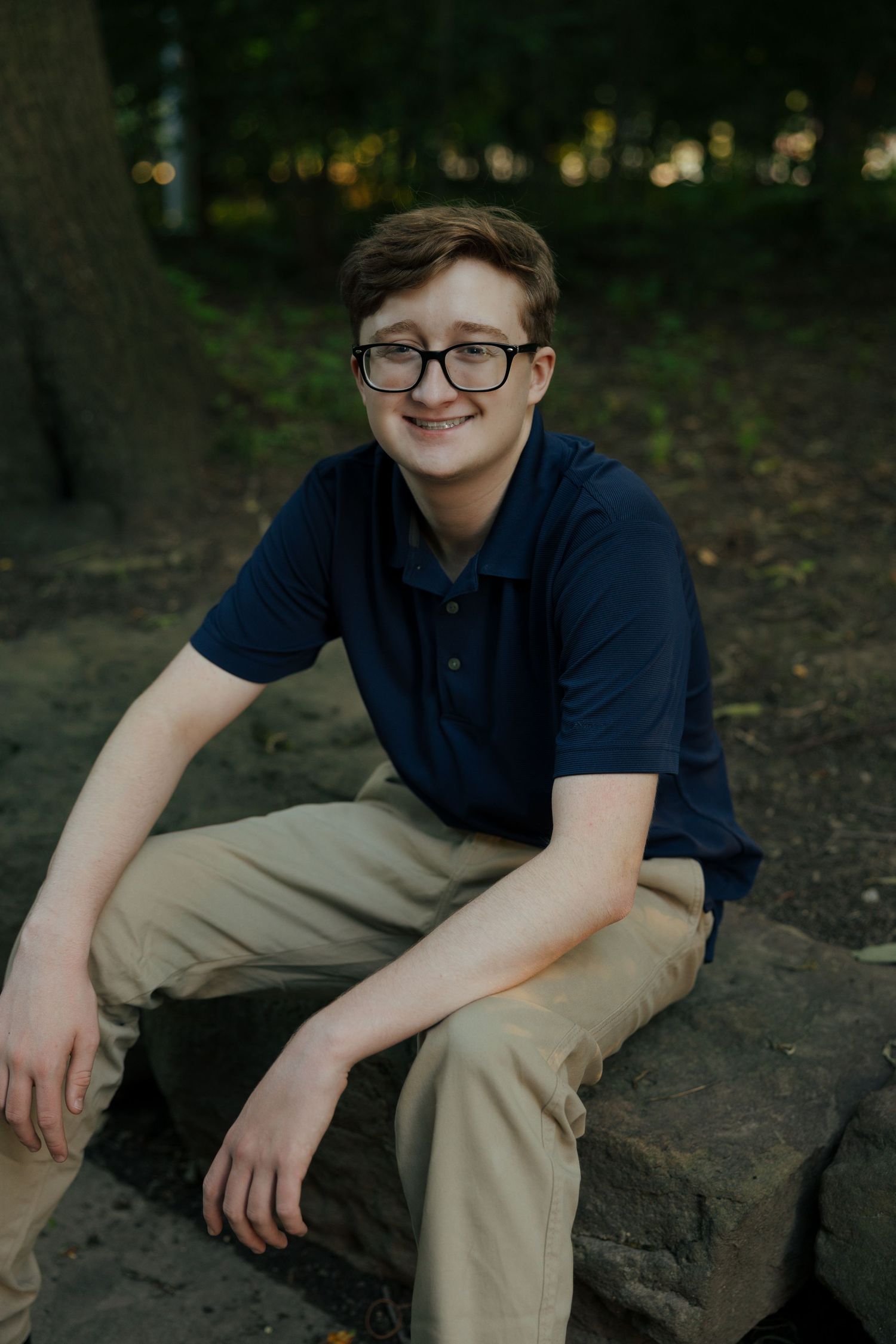 Smiling teenage boy with glasses and braces sits on stone wall in wooded park setting.