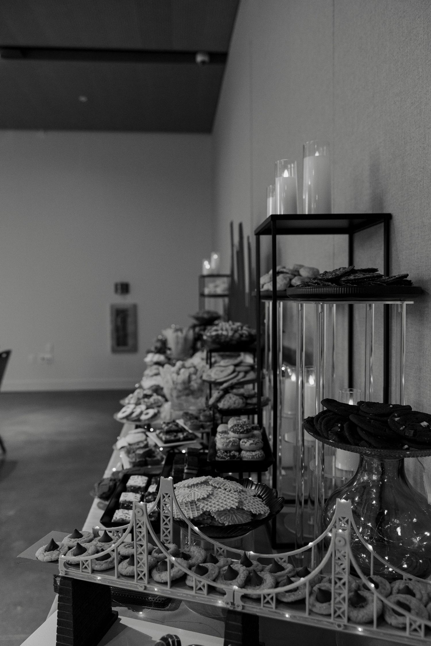 Black and white photo of an elegant dessert table display with candles, tiered stands, and assorted sweets at an event.
