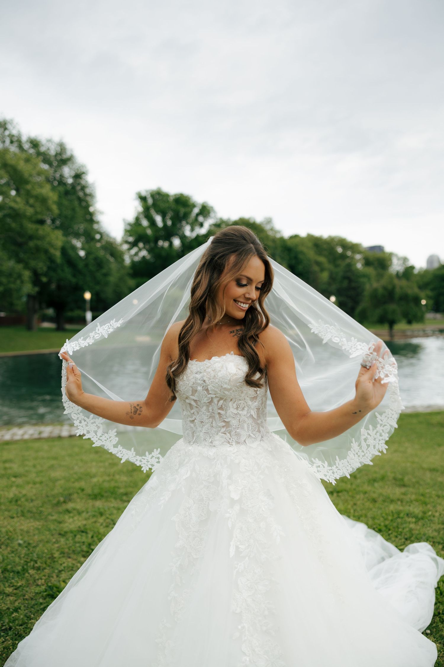 Smiling bride in strapless lace ball gown holding veil outdoors near pond with green trees.