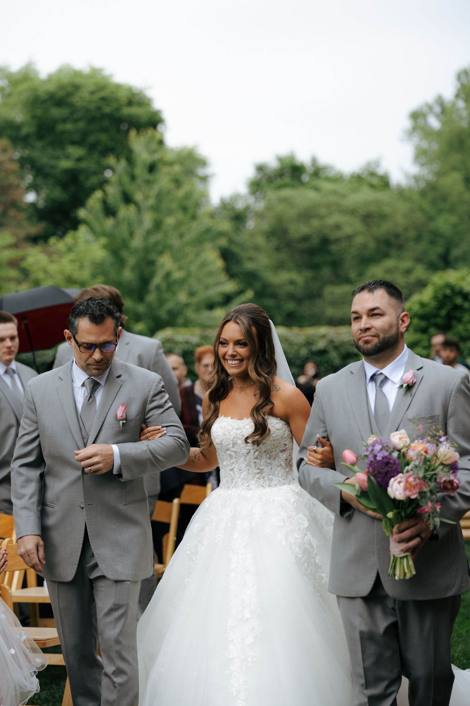 Smiling bride walks down outdoor aisle in lace ball gown, escorted by two men in gray suits holding bouquet.