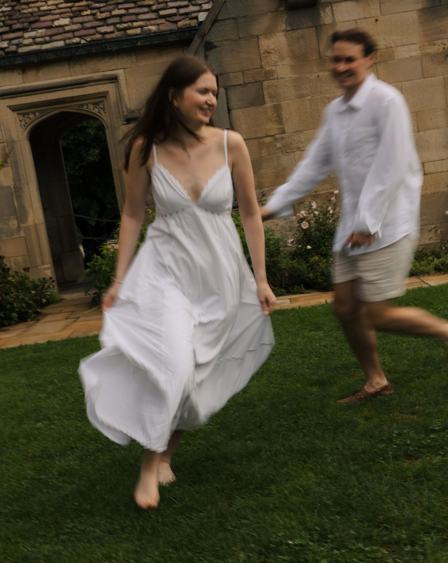 Woman in flowing white dress running barefoot on grass with man in white shirt at estate.