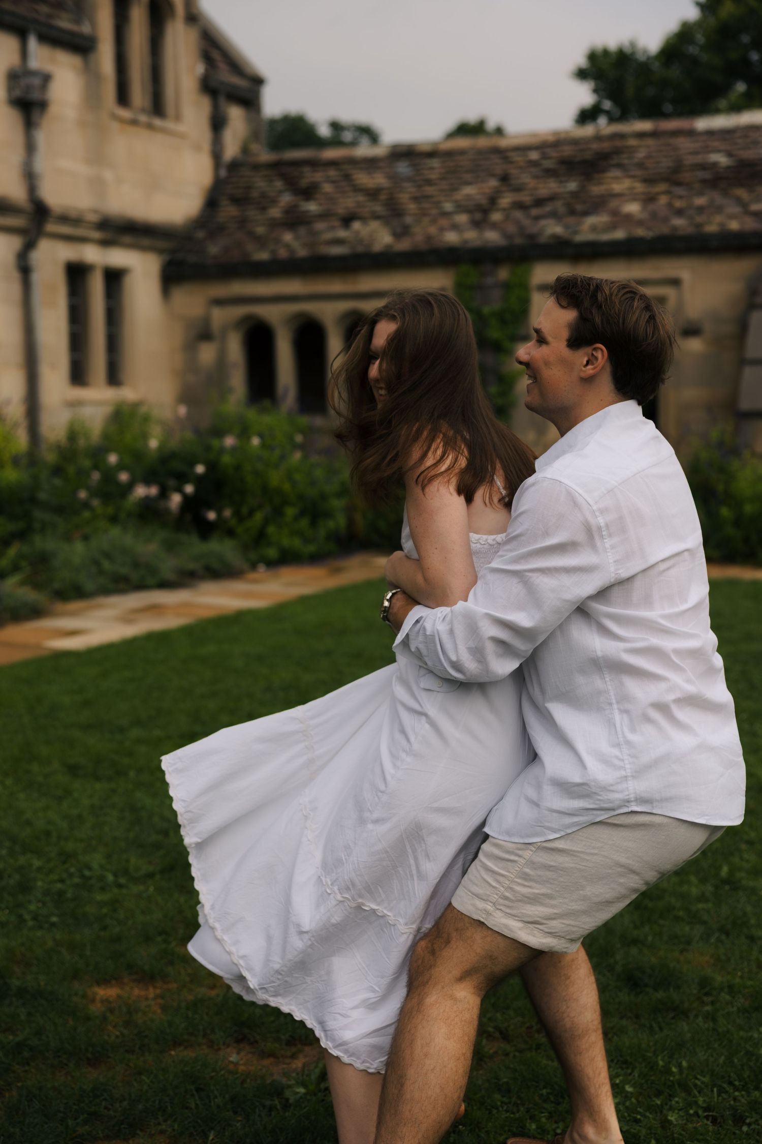 Couple in white outfits dancing and spinning together on a lush green lawn by a stone estate.