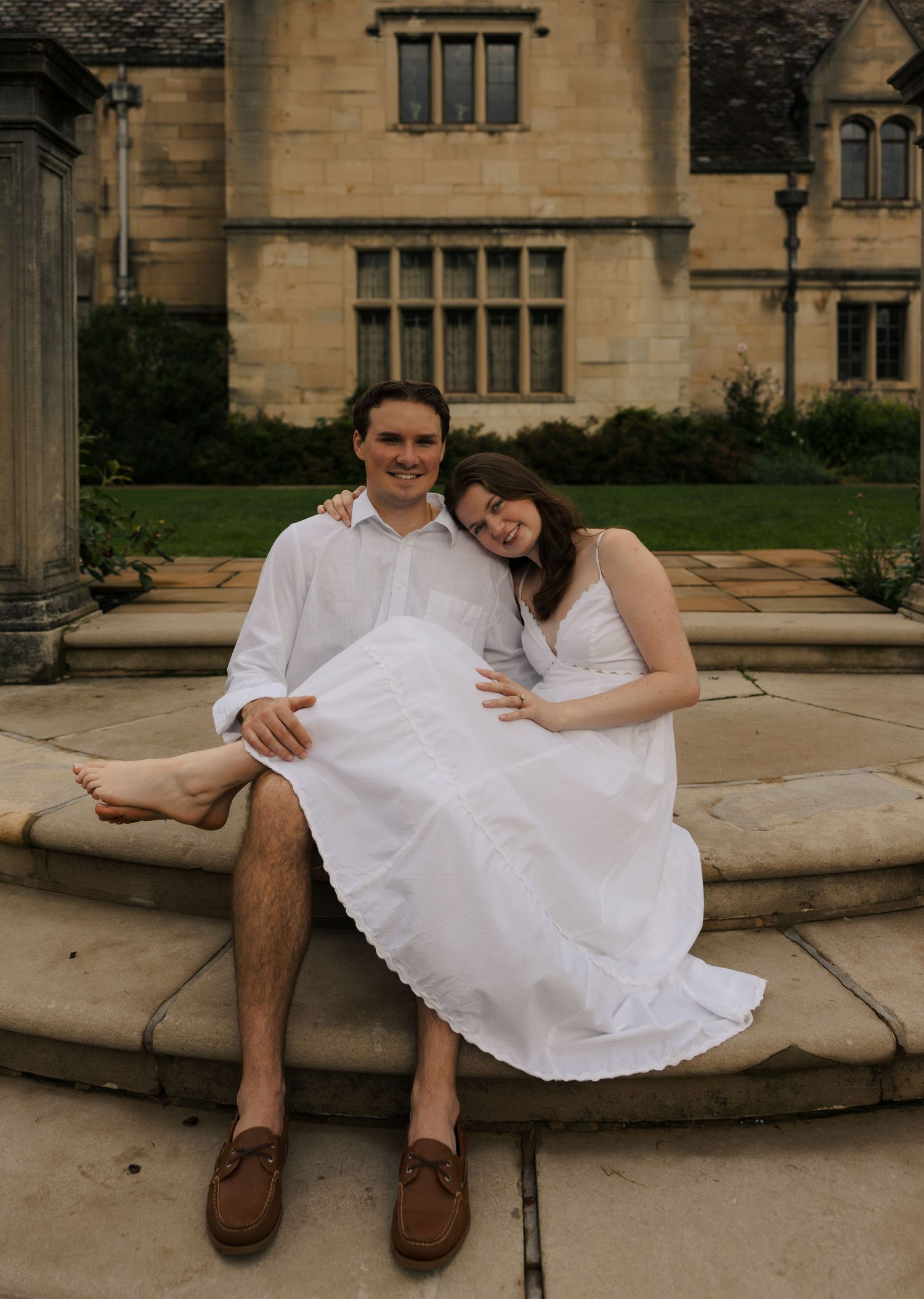 A smiling couple in white outfits sits relaxed on stone steps in front of a classic English manor house.