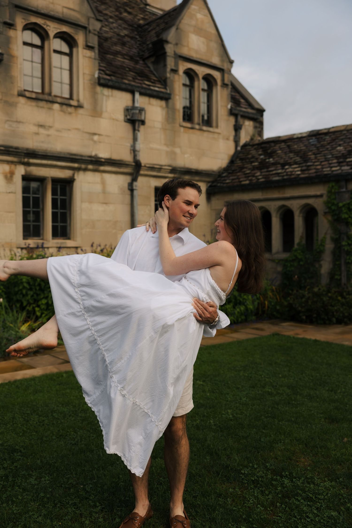 Man carrying woman in white dress while gazing lovingly at each other on green lawn.