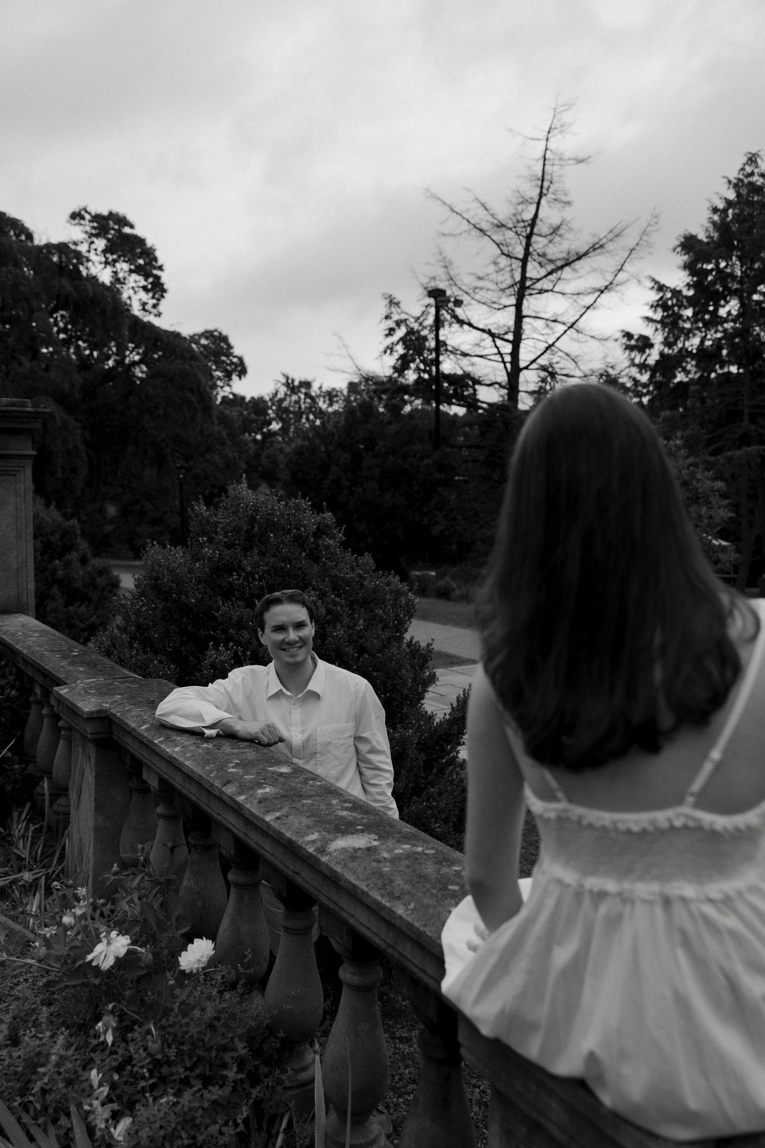 Black and white photo of a smiling man leaning on a stone railing as a woman in white approaches him.