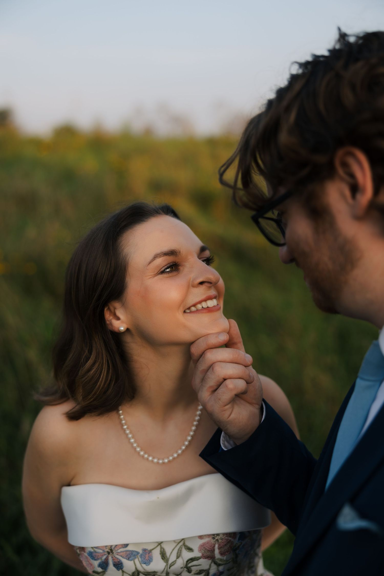 A smiling bride in a white strapless gown and pearl necklace gazes up at a groom in a blue suit outdoors.