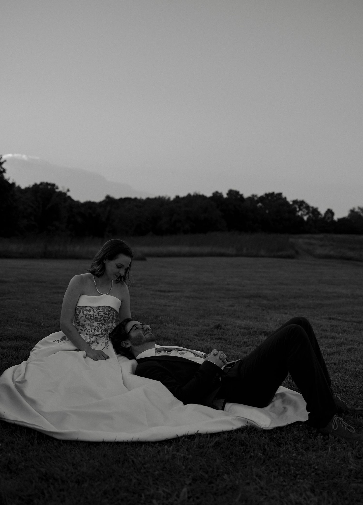 Black and white photo of bride in strapless gown with groom resting on her lap in an open field.