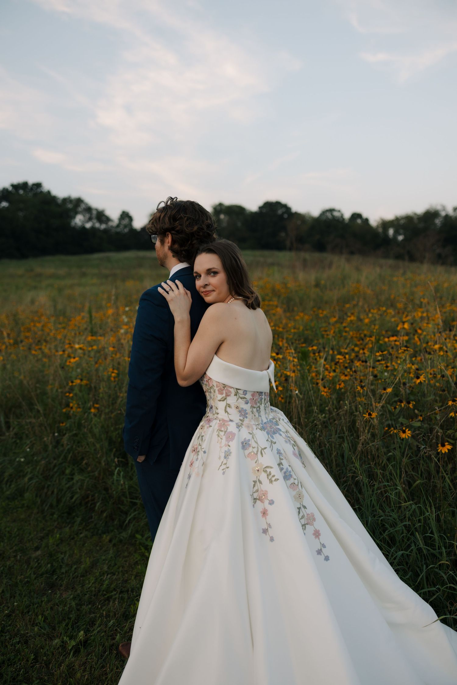Bride in floral off-shoulder ball gown embraces groom from behind in vibrant yellow wildflower field.