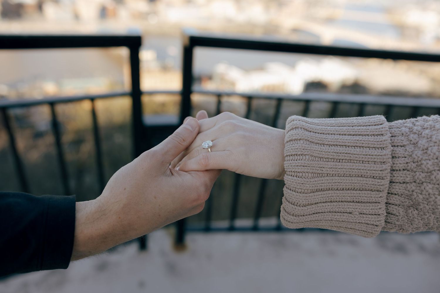 A hand in a gray sweater sleeve reaches out against a blurred background of a balcony railing and city view.