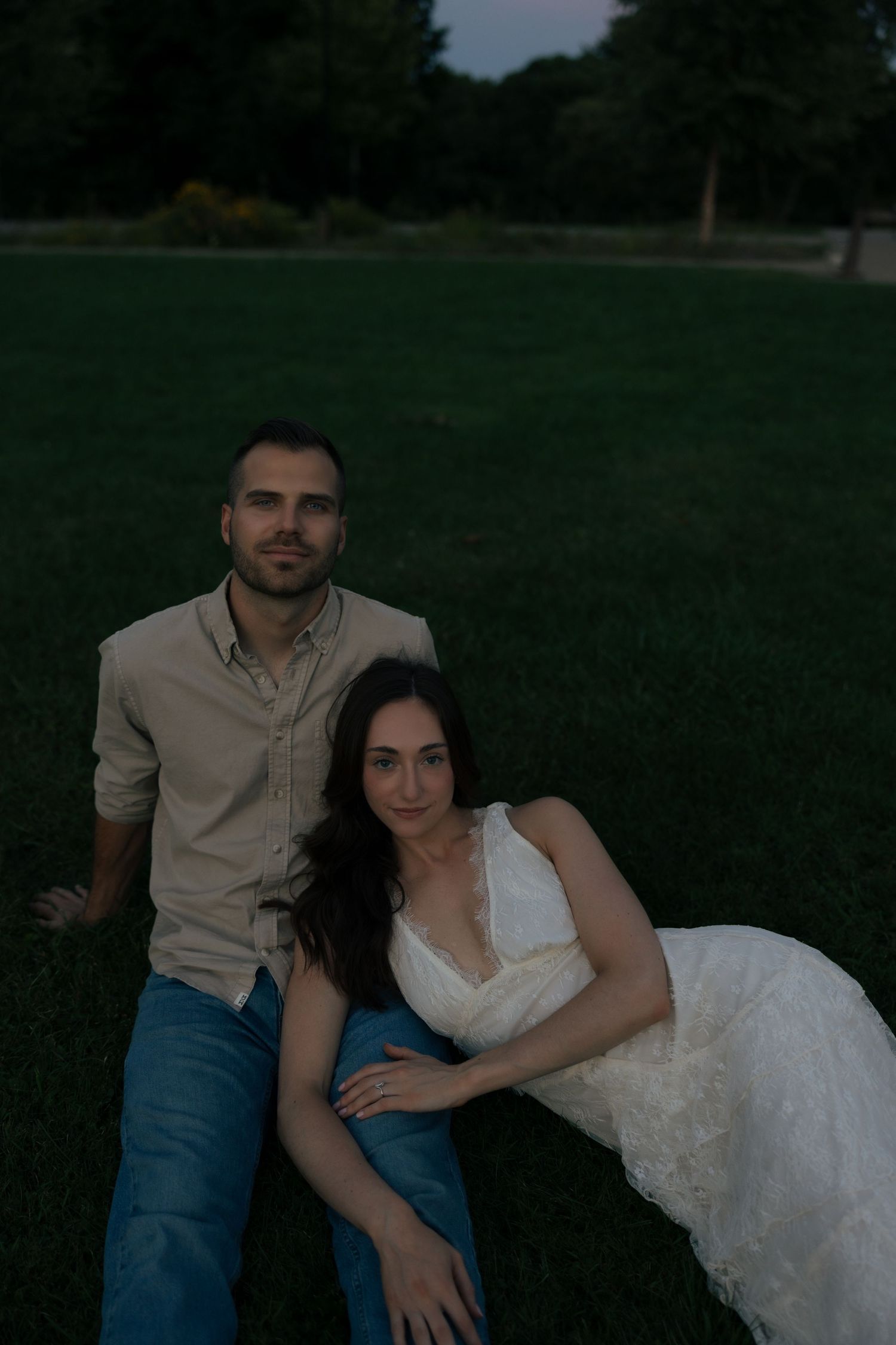 A couple sits together on lush green grass at dusk, woman in white dress leaning against man in beige shirt and jeans.