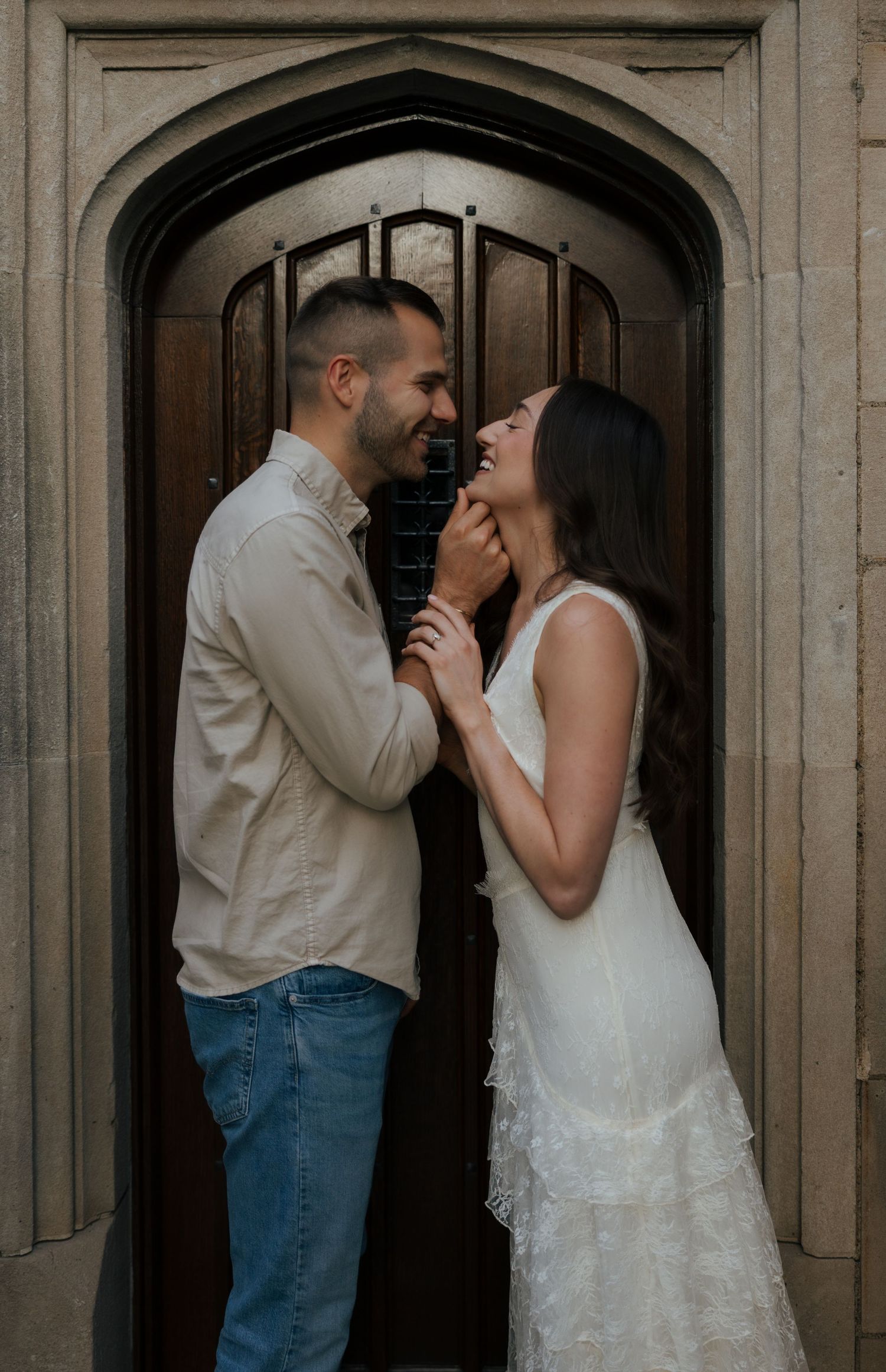A couple shares a joyful moment in front of an arched wooden door, the man gently touching the woman's face as she laughs.