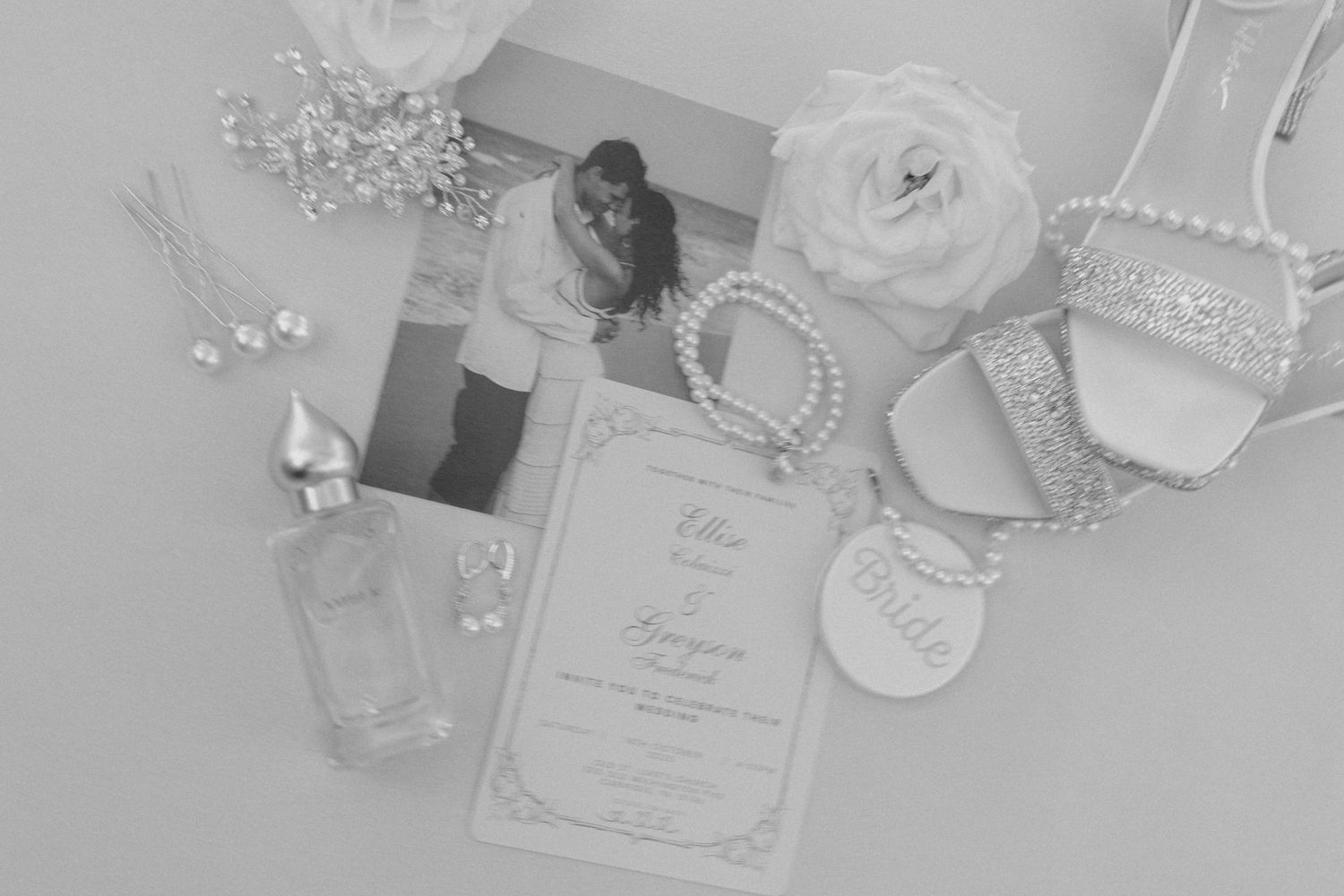 Black and white flat lay of wedding stationery, pearl bracelet, heels, perfume, floral accessories, and a couple photo.