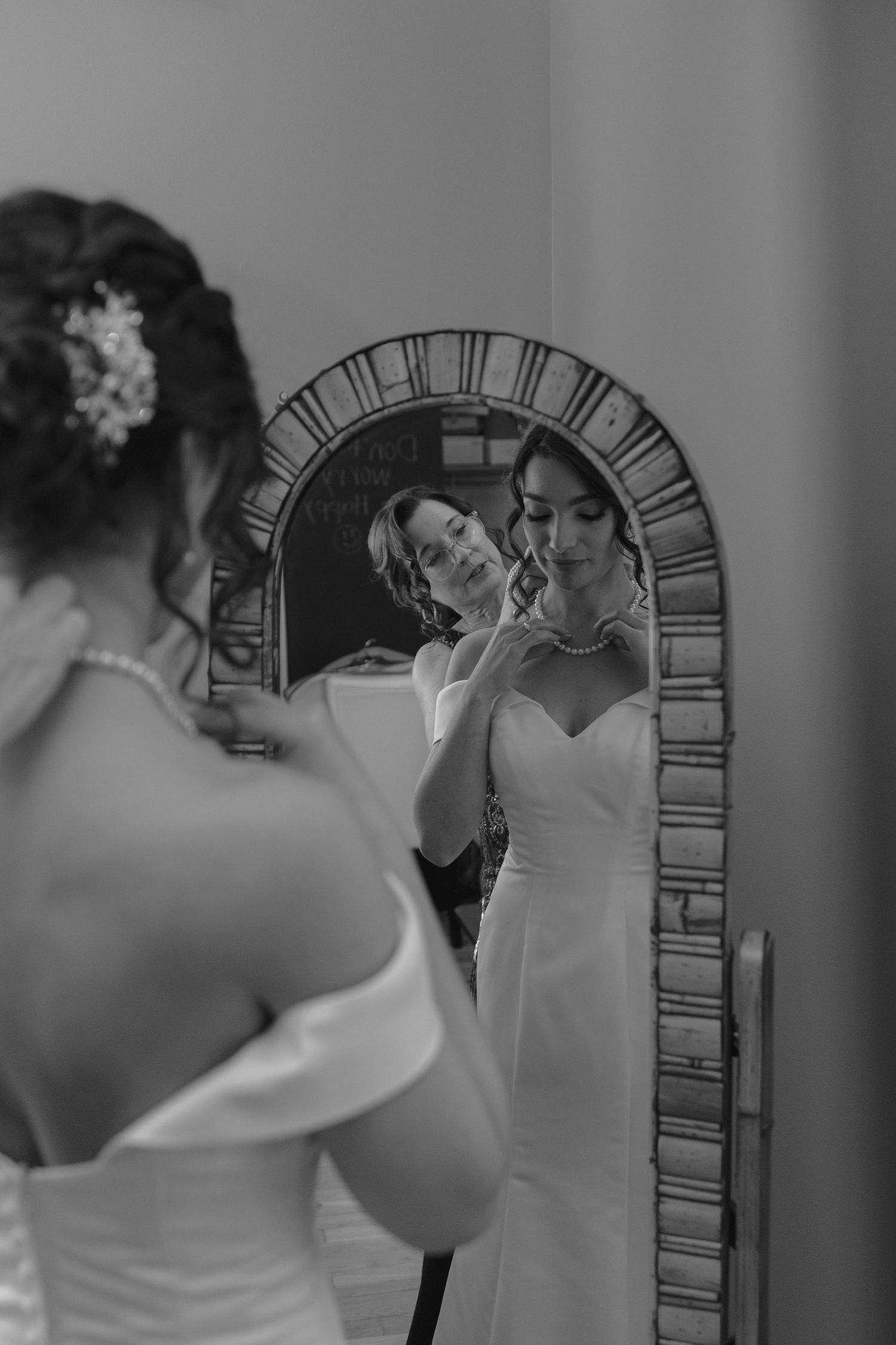 Black and white photo of a bride in a white gown adjusting her necklace while looking into an ornate arched mirror.