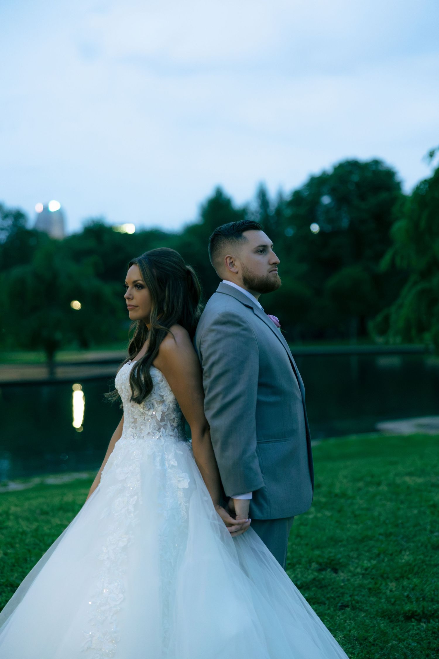 Bride and groom stand back-to-back holding hands in a lush park at dusk, bride in white gown, groom in gray suit.