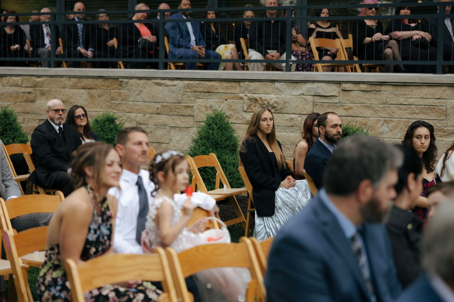 Wedding guests seated in wooden chairs at outdoor ceremony with upper-level spectators watching above.