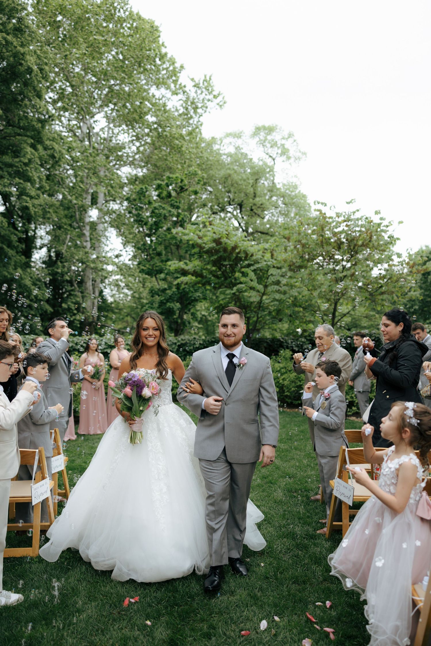 Newlyweds walk down the aisle as guests blow bubbles during outdoor garden wedding ceremony.