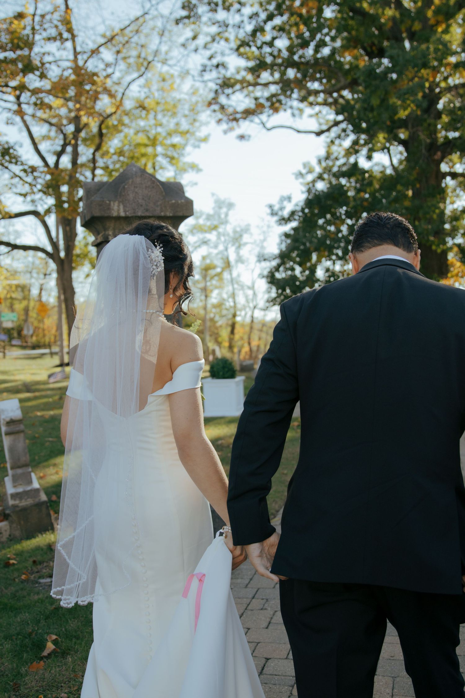 Bride in white gown and veil walks hand in hand with groom through an outdoor garden on their wedding day.